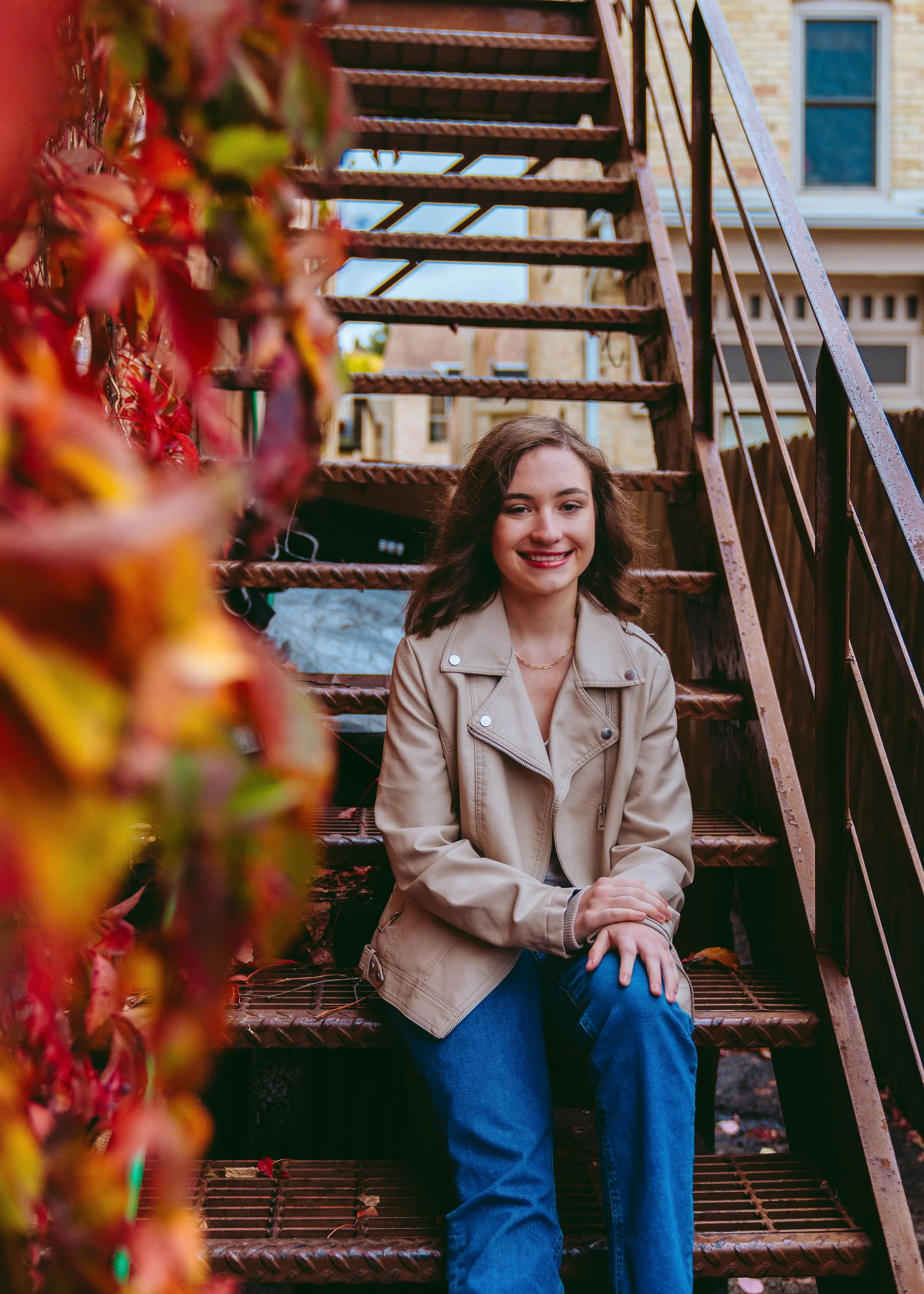 A young woman with shoulder-length brown hair, smiling, sitting on outdoor rusted metal stairs with a beige jacket, blue jeans, and a gold necklace, framed by colorful autumn leaves on the left, with brick buildings in the background.