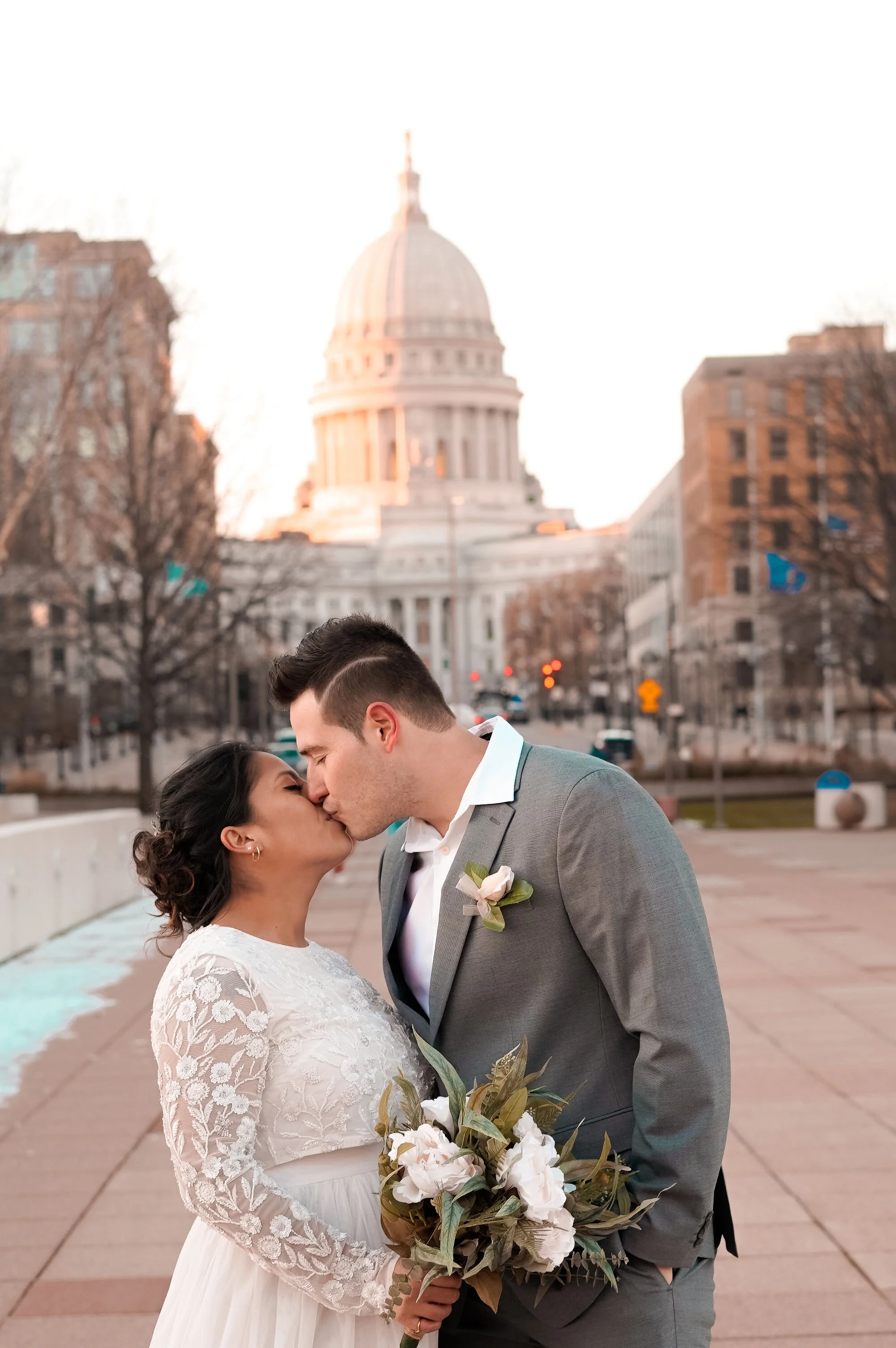A couple in wedding attire sharing a kiss in front of the United States Capitol building during sunset.