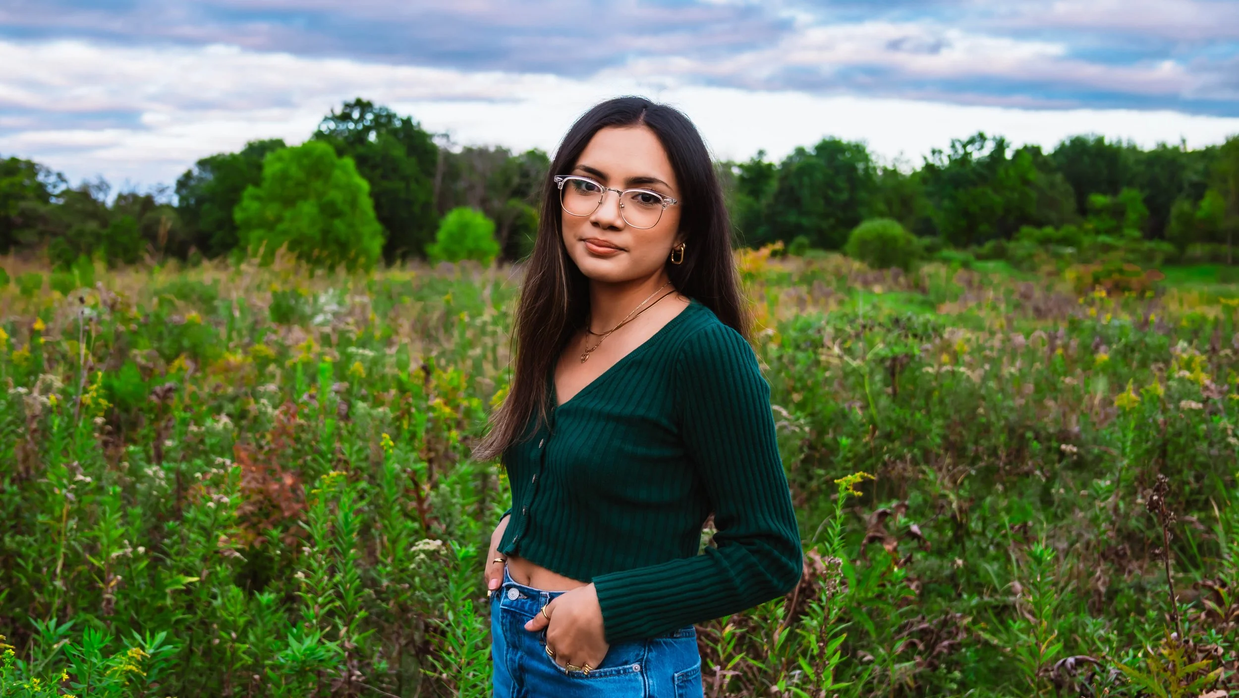 Young woman with long dark hair, glasses, and gold jewelry stands in a lush green field with trees and a cloudy sky in the background.