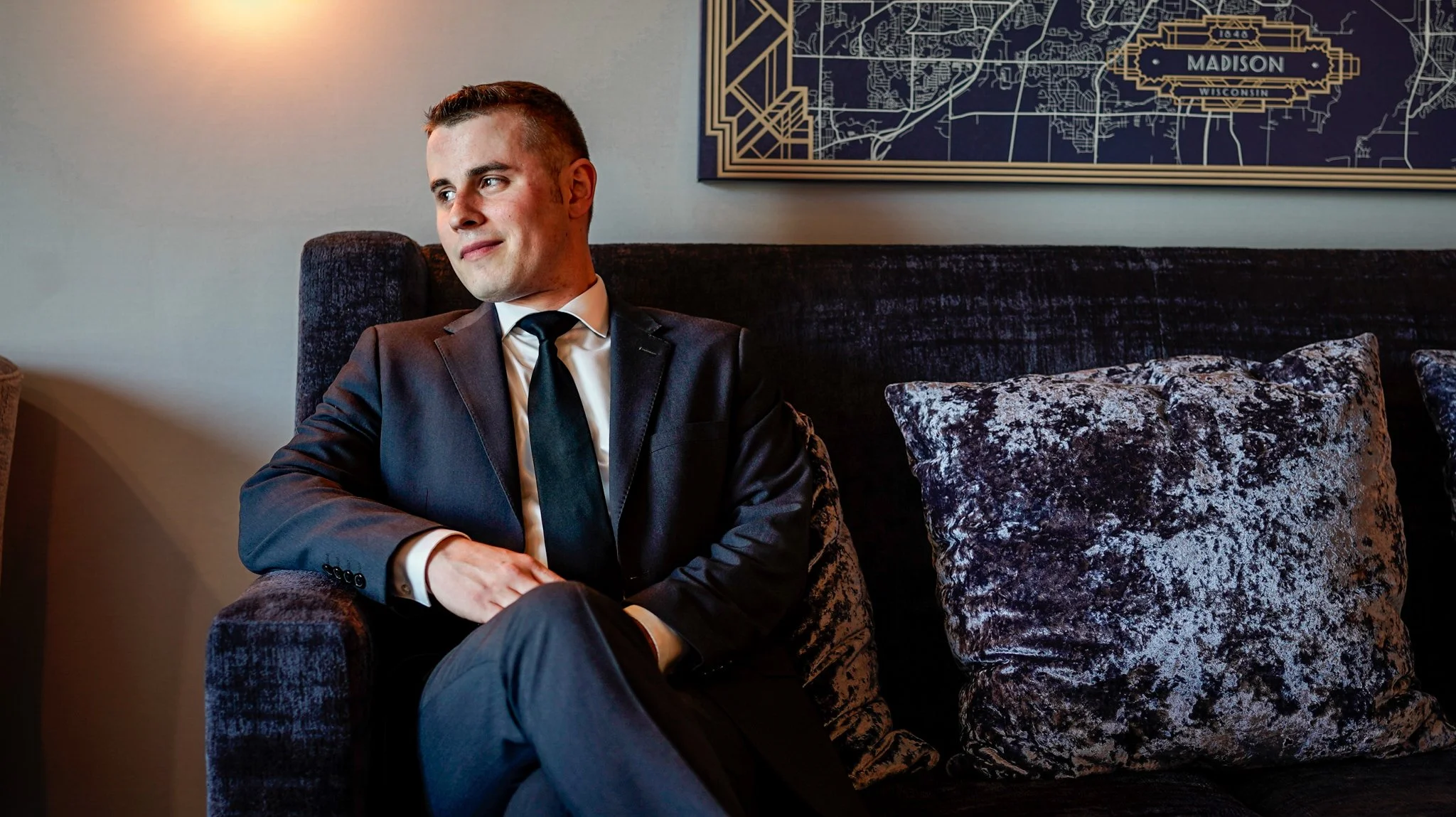 A man in a black suit and tie sitting on a dark velvet sofa with patterned pillows, looking thoughtfully to the side, with a framed map of Madison, Wisconsin hanging on the wall behind him.