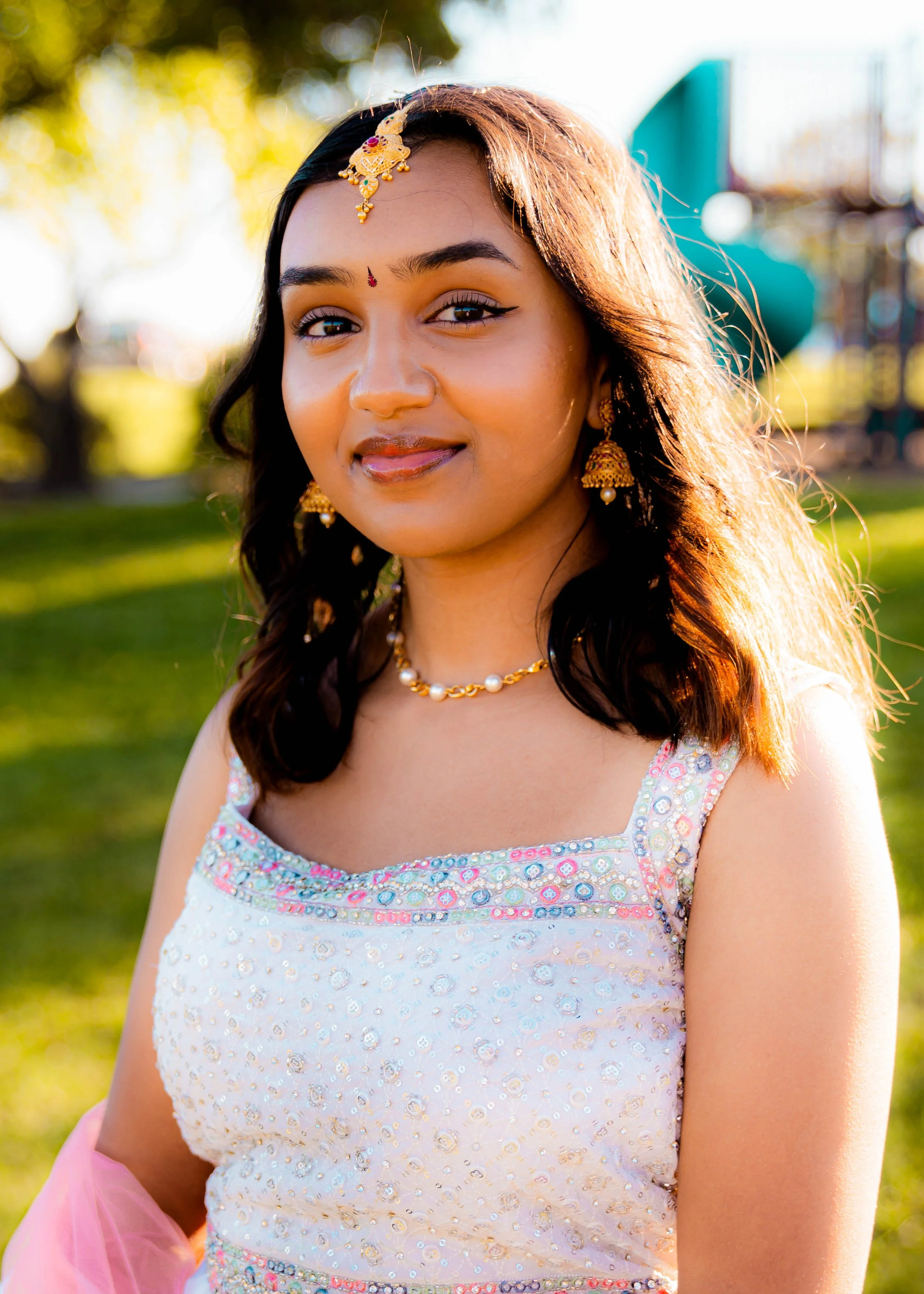 A woman outdoors in sunlight, wearing traditional Indian jewelry and attire, with a playground slide visible in the background.
