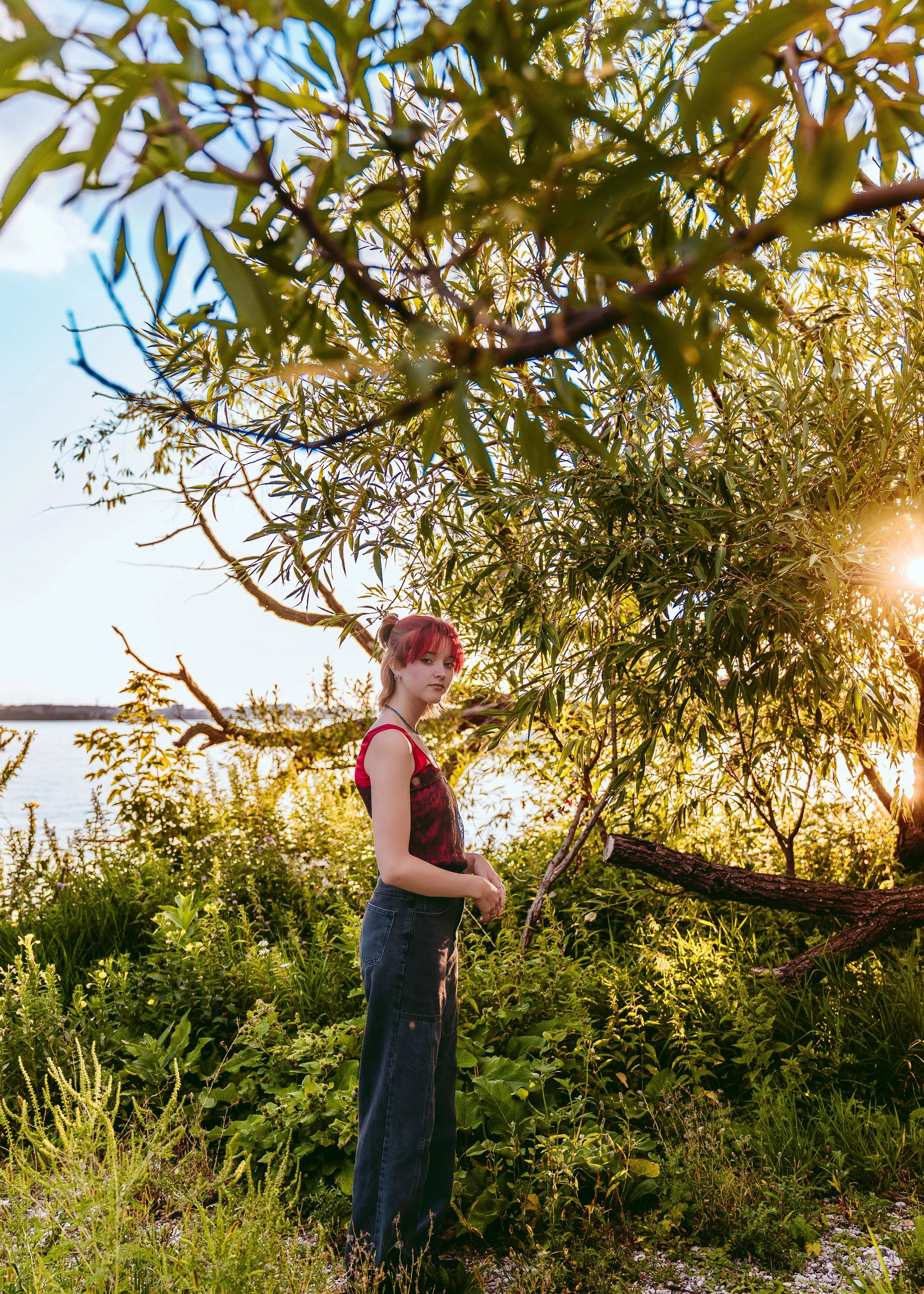 A young woman with red hair and a black and red sleeveless top standing outdoors among green trees and plants near a body of water during sunset.