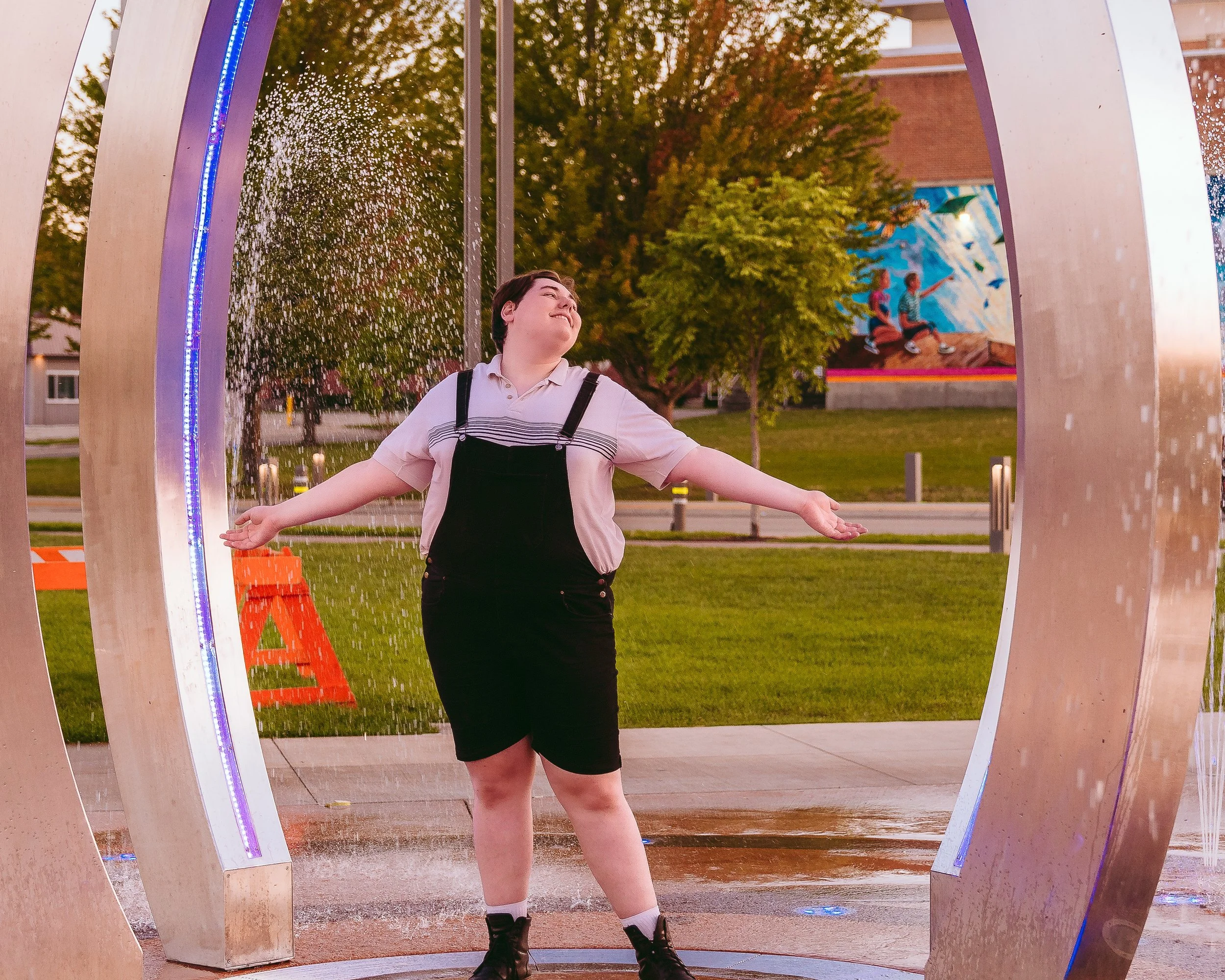 A person standing with arms outstretched inside a circular water fountain at sunset in a park, with trees, a mural wall, and orange traffic cones visible in the background.