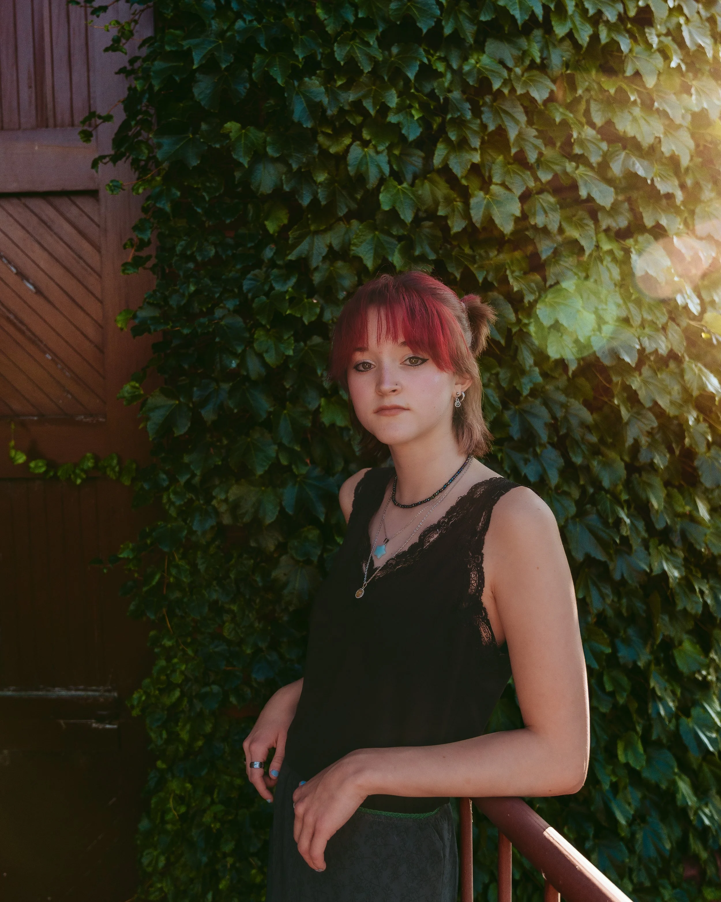A young woman with short red hair and bangs, wearing a black sleeveless lace top, stands outdoors in front of a lush green ivy-covered wall, with sunlight filtering in on the right side.