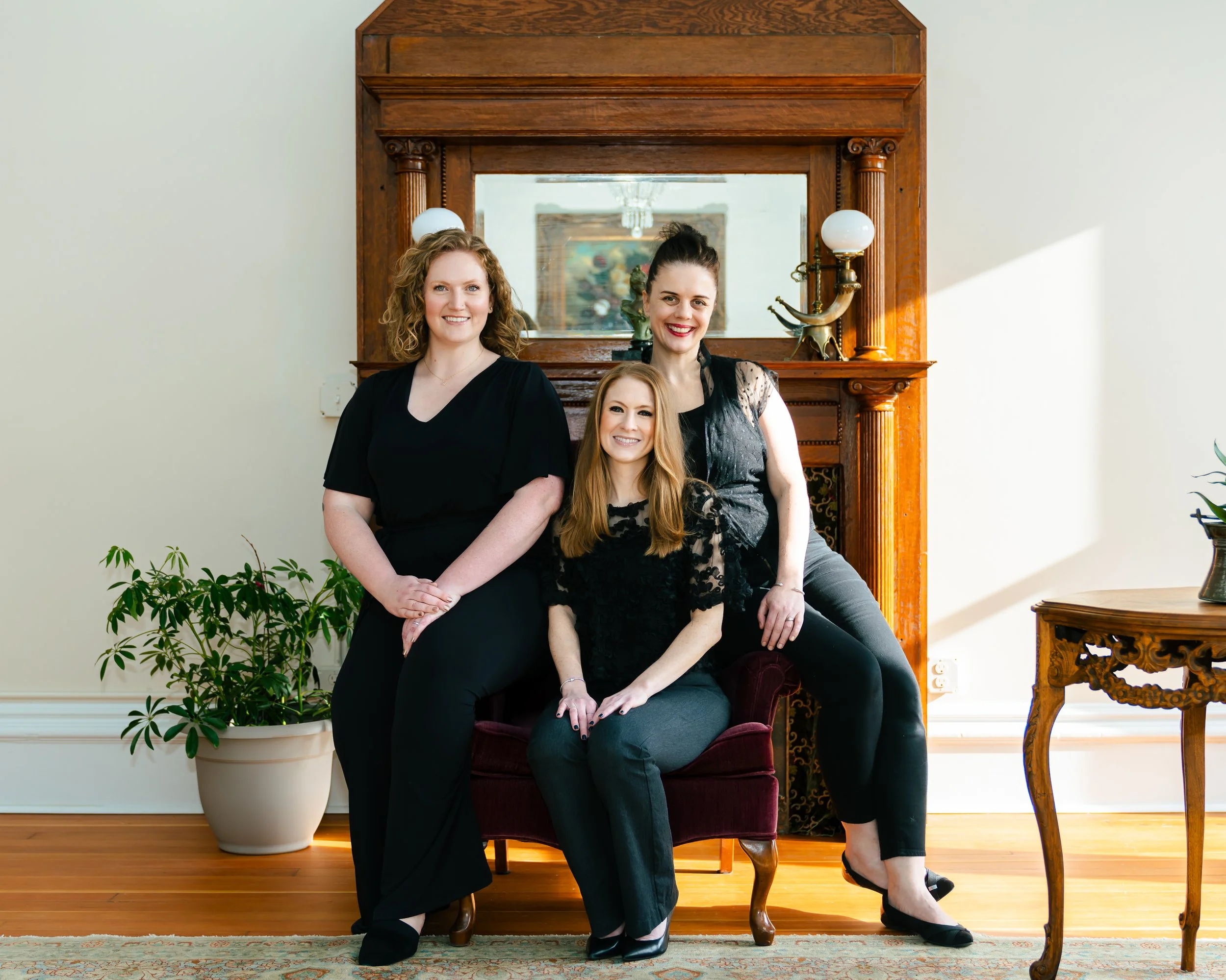 Three women in black outfits smiling and posing in a living room with a wooden mirror, plants, and antique furniture.