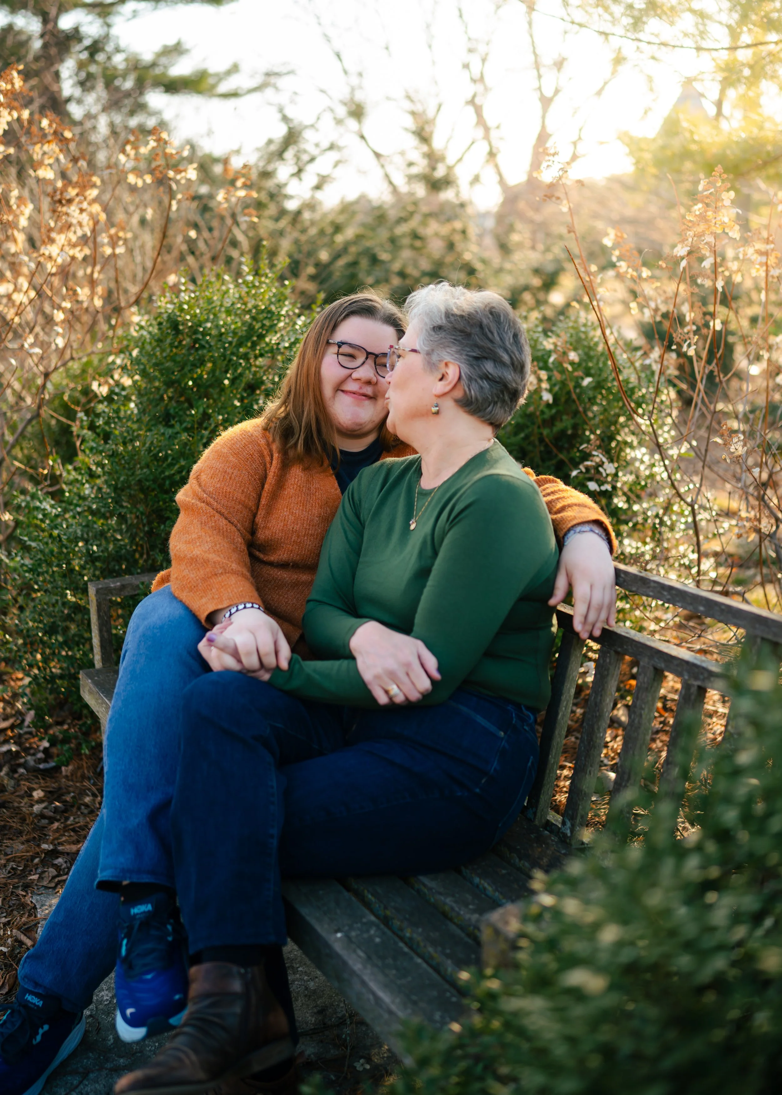 A woman and a teenage girl share a tender moment on a park bench surrounded by greenery and blooming trees in warm sunlight.