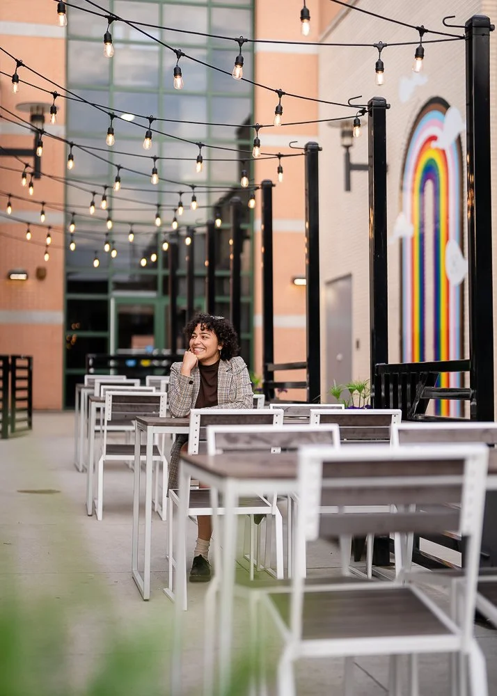 A woman sitting alone at an outdoor cafe with string lights overhead, brick buildings, and a colorful rainbow mural on a wall in the background.