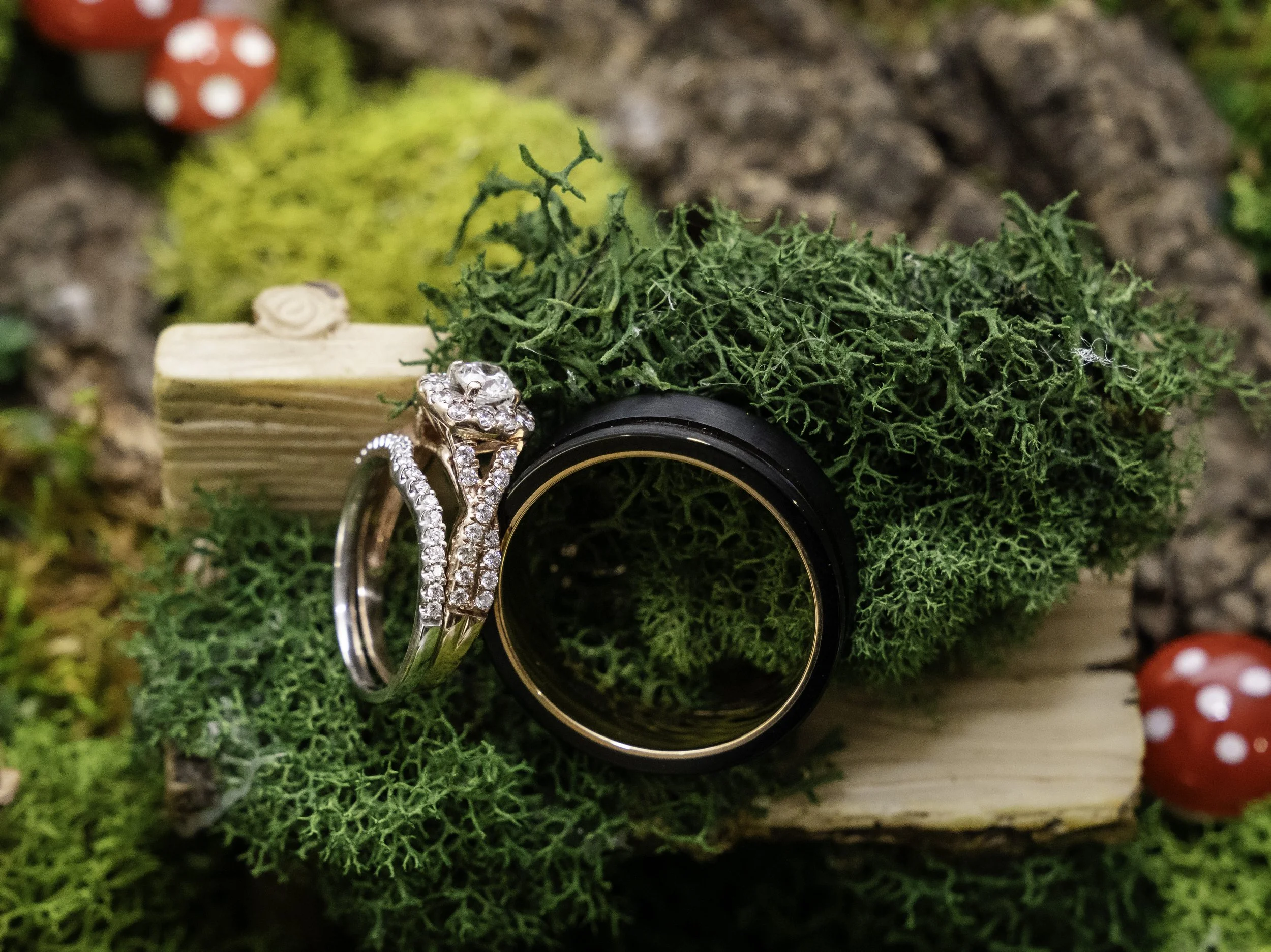 Close-up of two wedding rings, one with diamonds and one plain, resting on a bed of green moss and foliage, with a small wooden block in the background and red and white mushroom decorations.