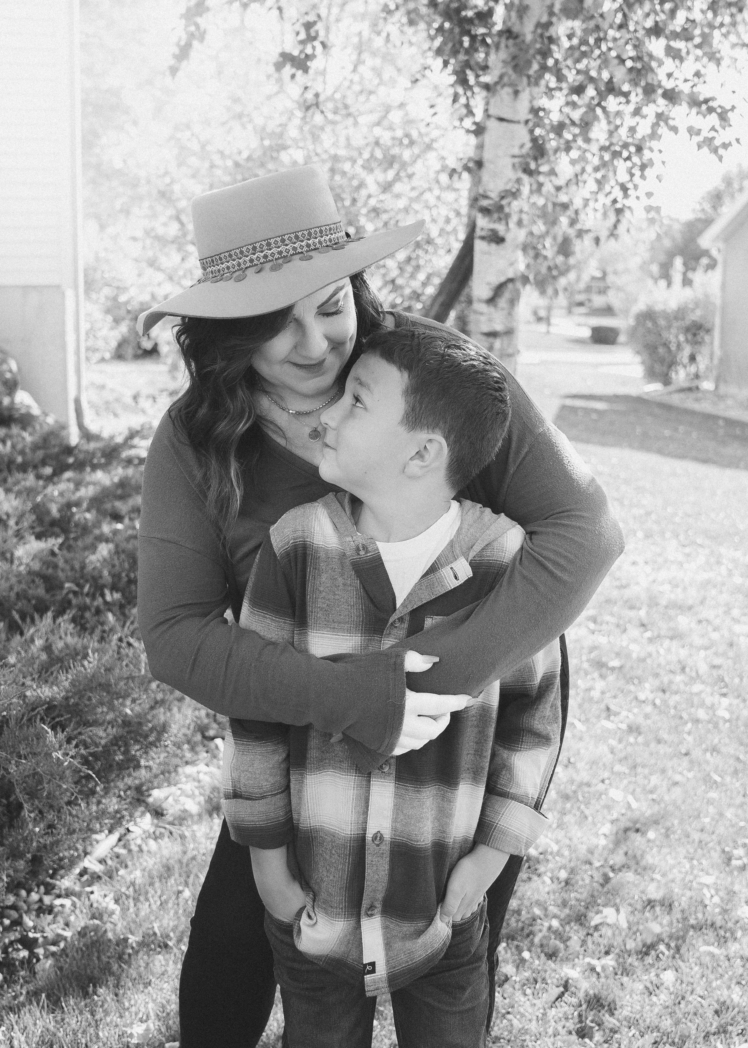 A woman wearing a wide-brimmed hat and a young boy are embracing outdoors, sharing a close moment with trees and a house in the background.