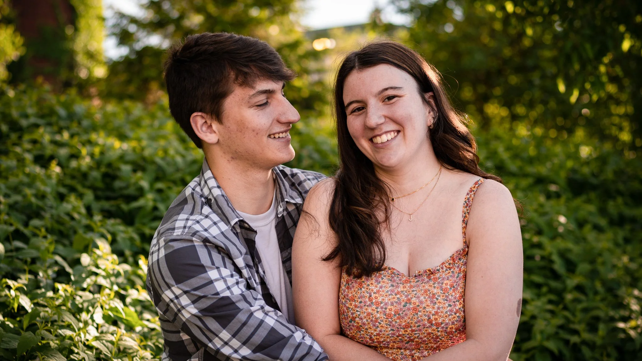 A young couple standing close together outdoors, smiling and looking at each other on a sunny day, with green foliage in the background.