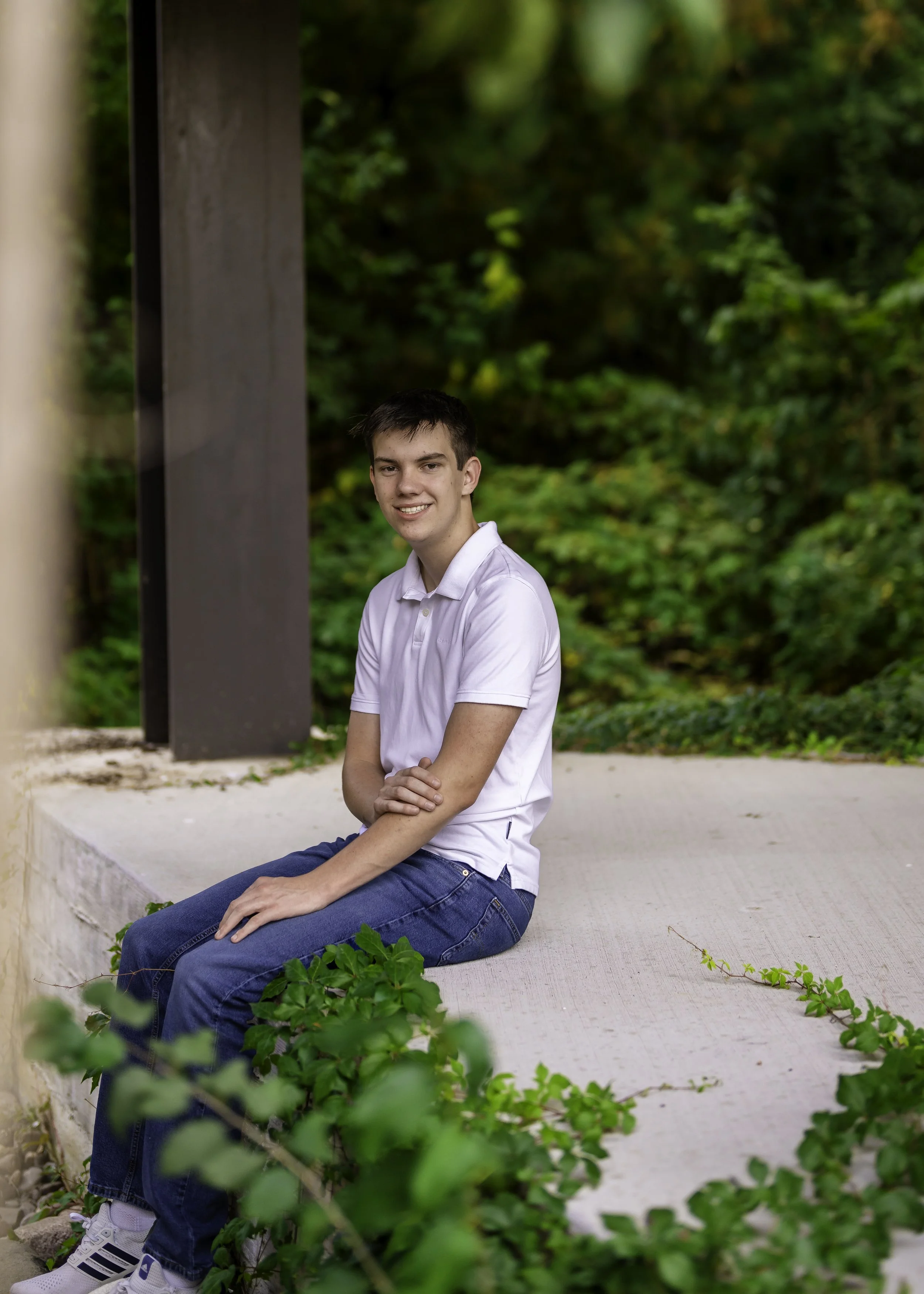 A young man sitting on a concrete ledge outdoors, wearing a white polo shirt and jeans, with green foliage in the background.