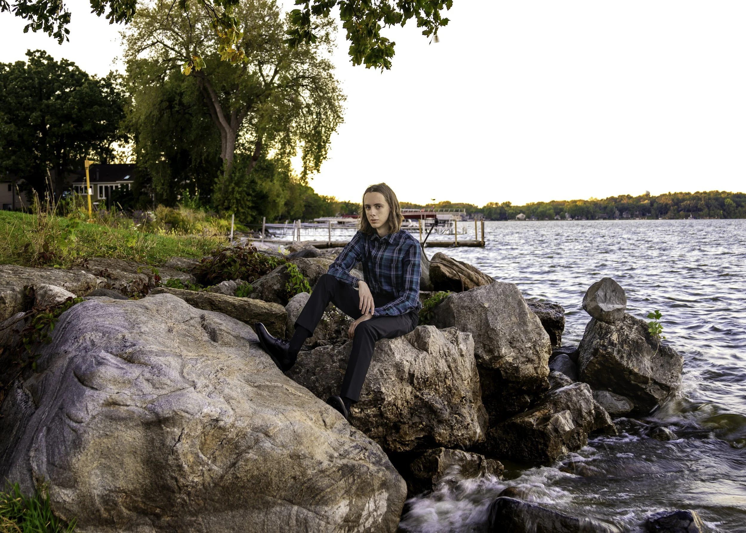 A woman sitting on large rocks at the edge of a lake during sunset, with trees and houses in the background.