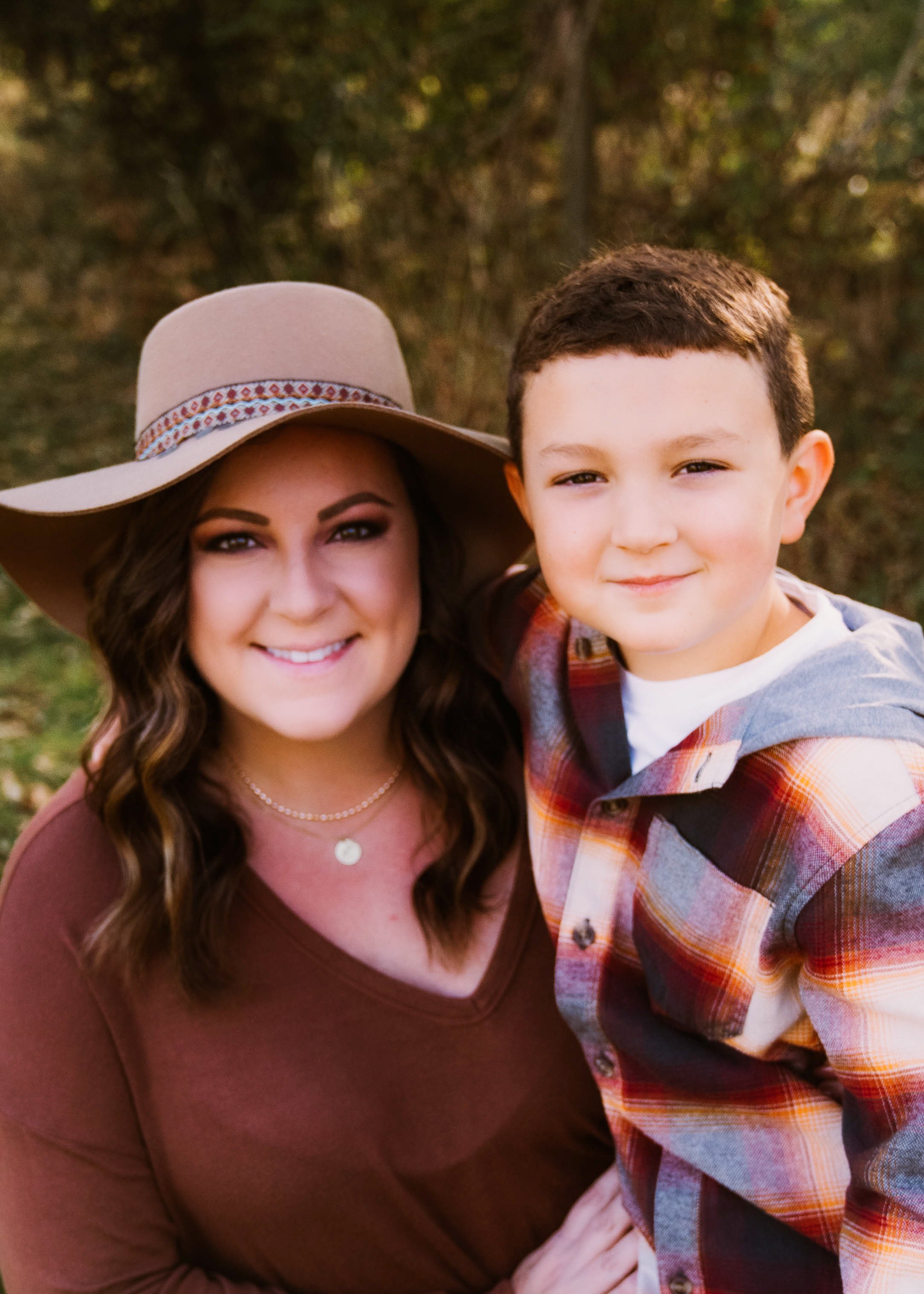 A woman wearing a large beige hat and a brown top, smiling, with a young boy in a plaid shirt, outdoors in a forested area.