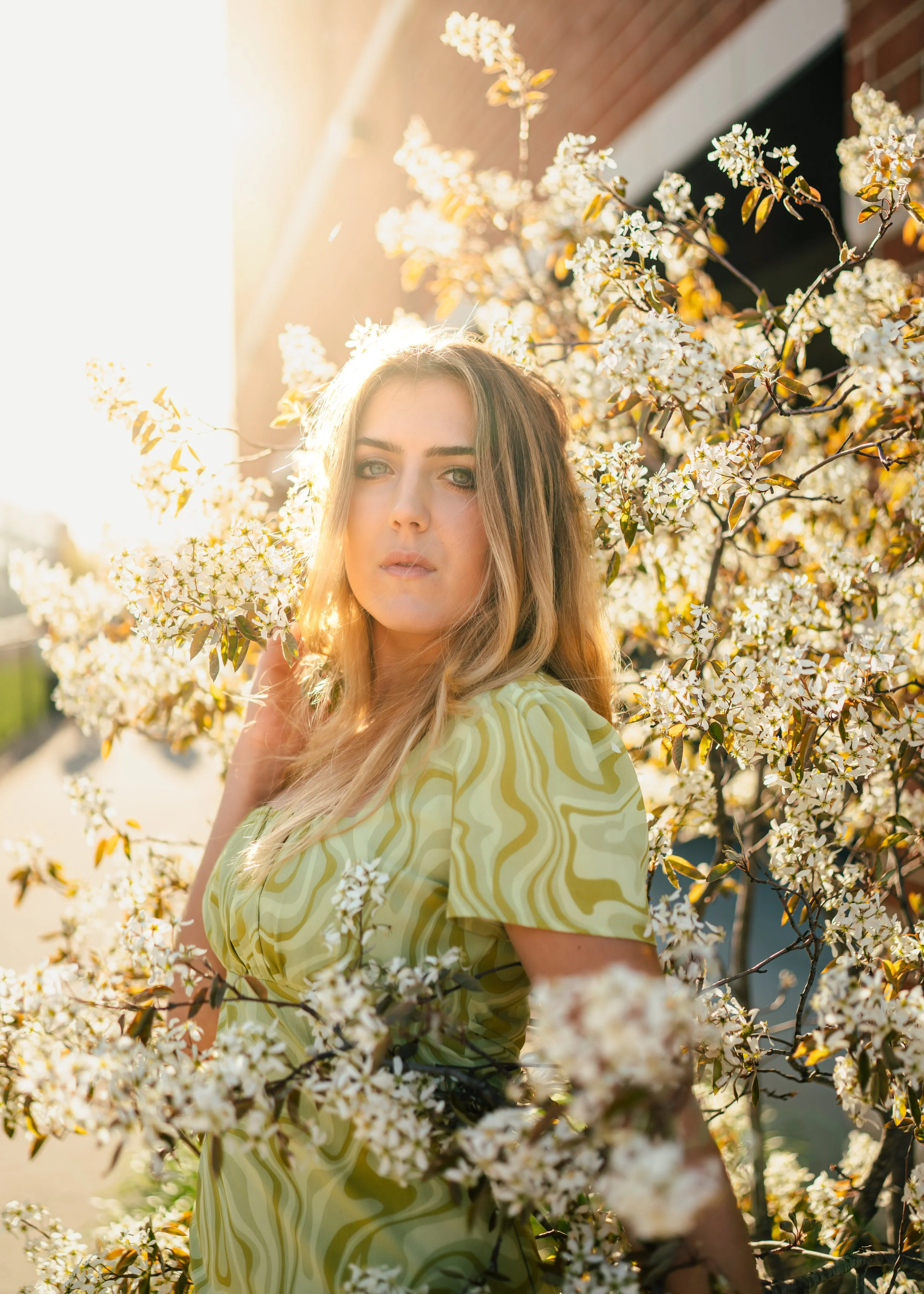A woman with long blonde hair and blue eyes posing among blooming white flowers on a tree, illuminated by warm sunlight.