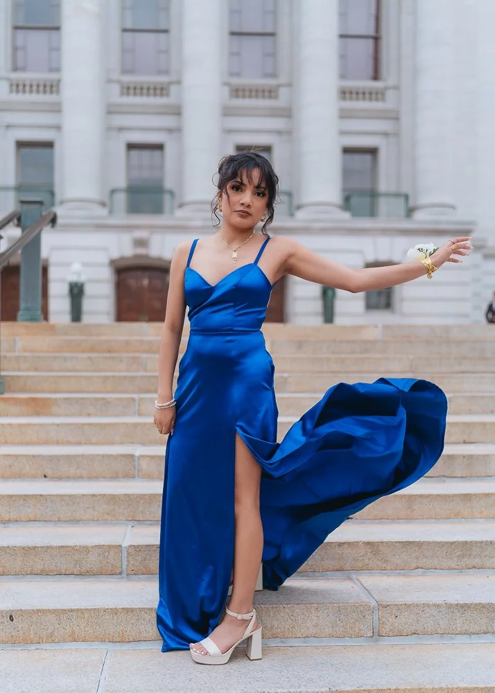 A woman in a blue satin evening gown with a high slit, standing on steps outside a large white building, holding one arm out to the side.