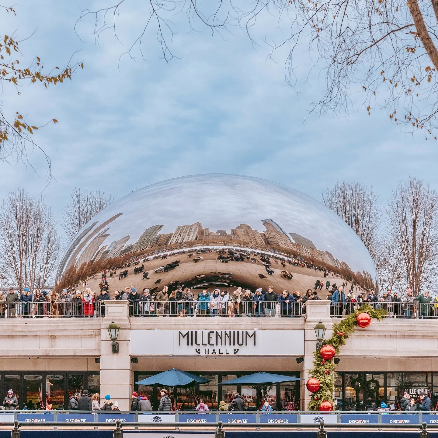 People gather outside Millennium Hall, a building with a large reflective sculpture on its roof, in a city setting during winter holidays, with some holiday decorations visible.