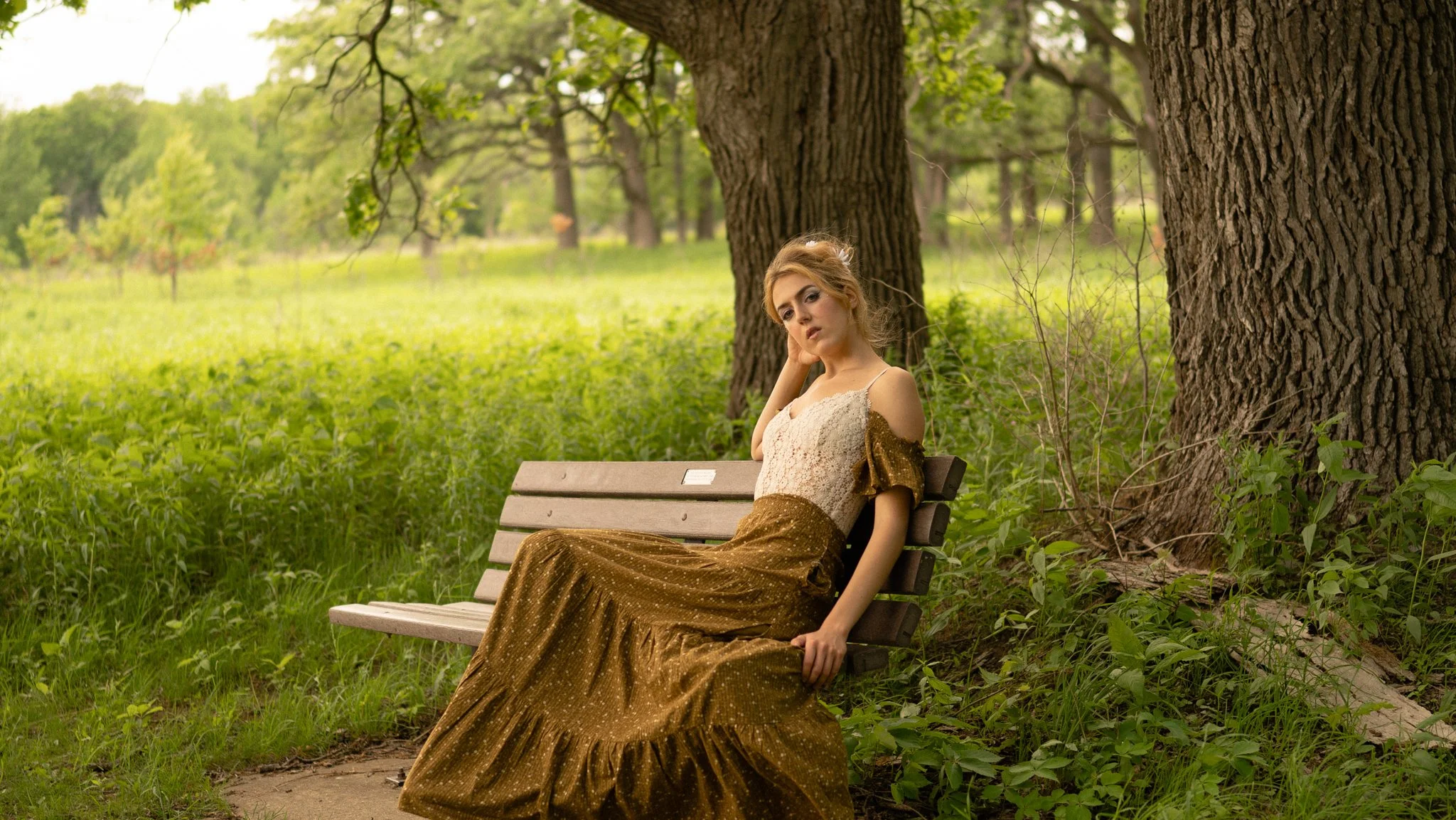 A woman in a vintage dress sitting on a park bench next to large trees and greenery, looking at the camera with a contemplative expression.