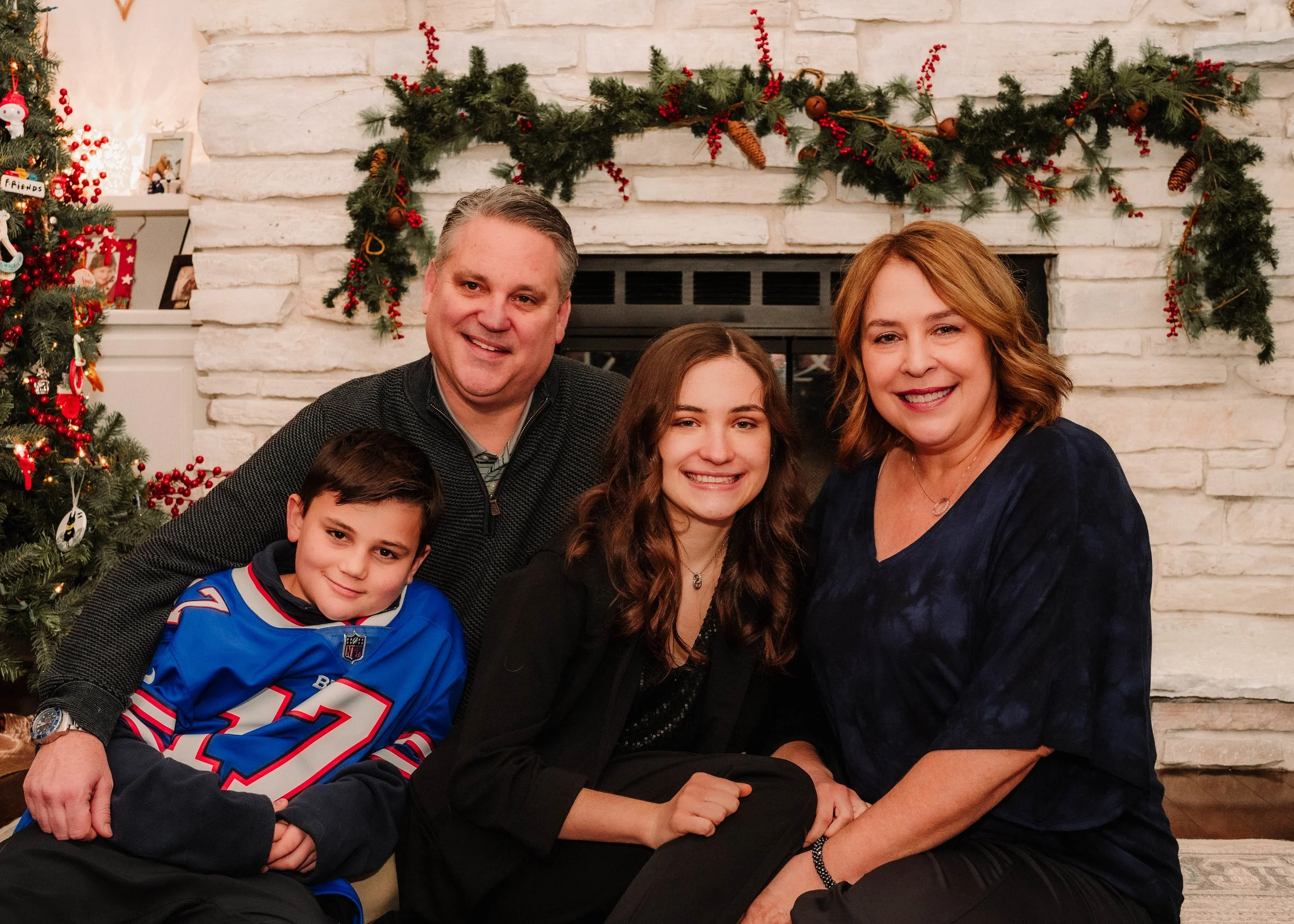 A family of four sitting in front of a fireplace decorated with Christmas garland and trees, smiling for a holiday photo.