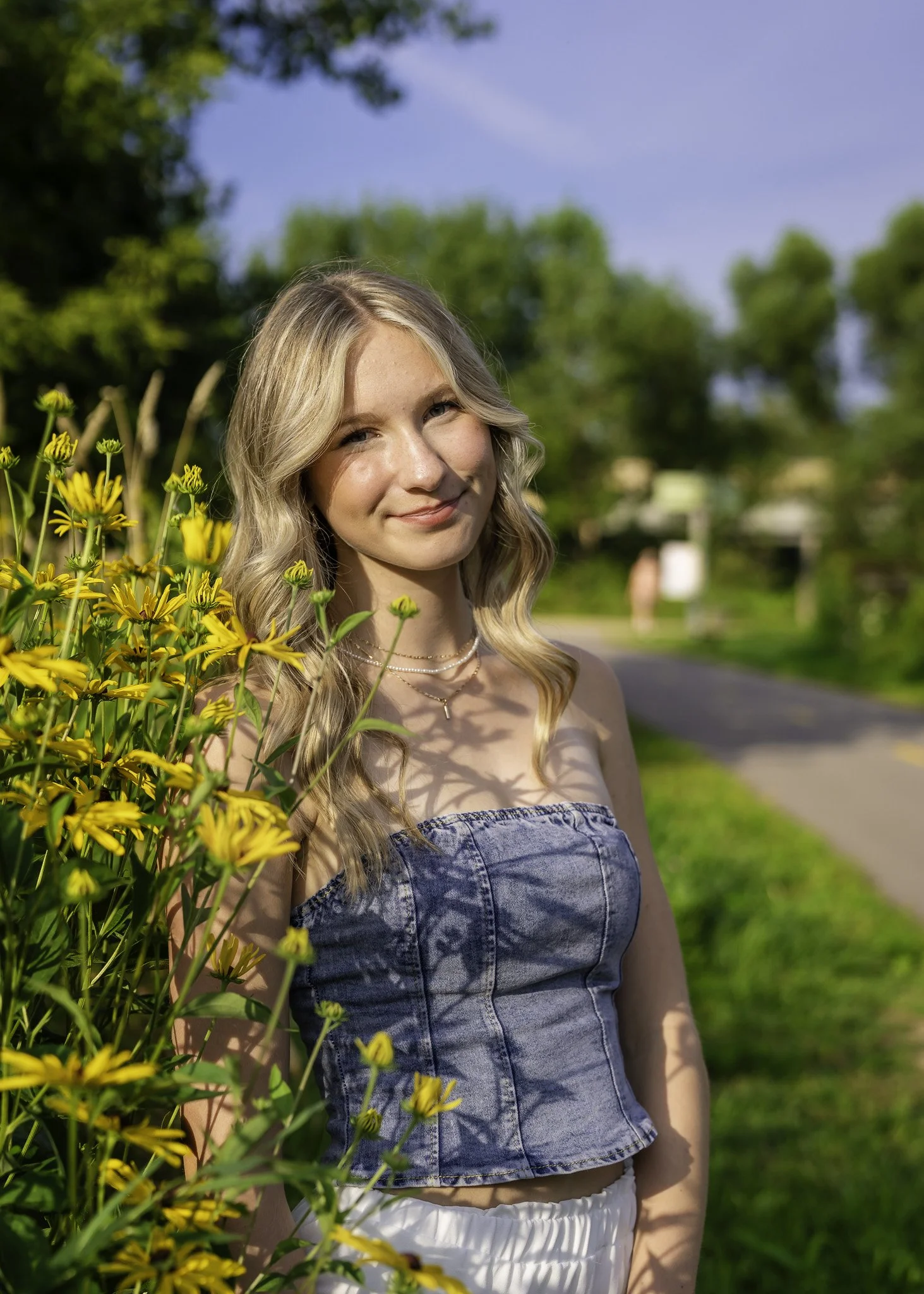 A young woman with long blonde hair and a denim tube top standing next to yellow flowers on a sunny day with a green landscape and trees in the background.