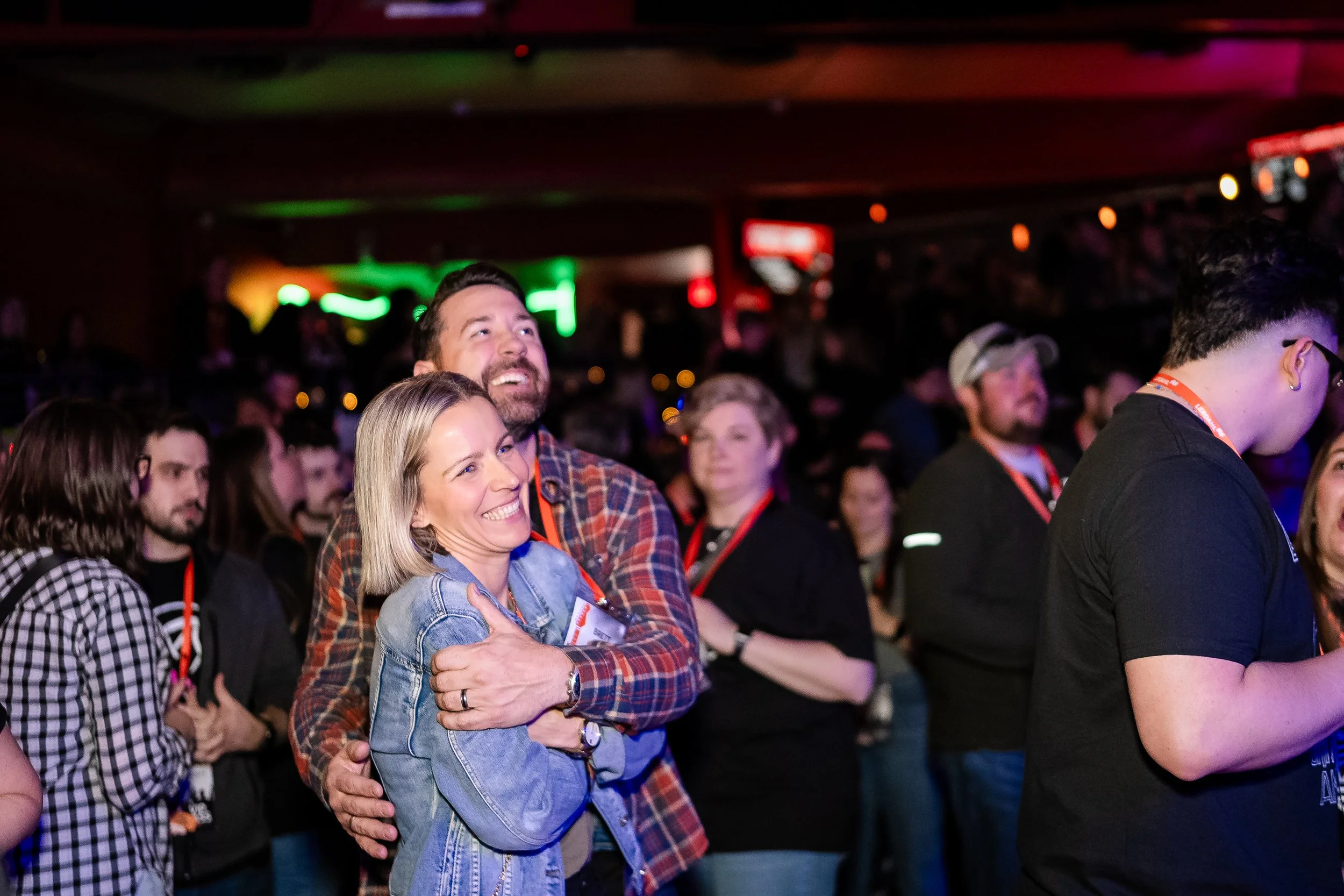 A crowded indoor event with people smiling and enjoying themselves, including a woman in a denim jacket and a man in a plaid shirt hugging, with colorful lighting reflected in the background.