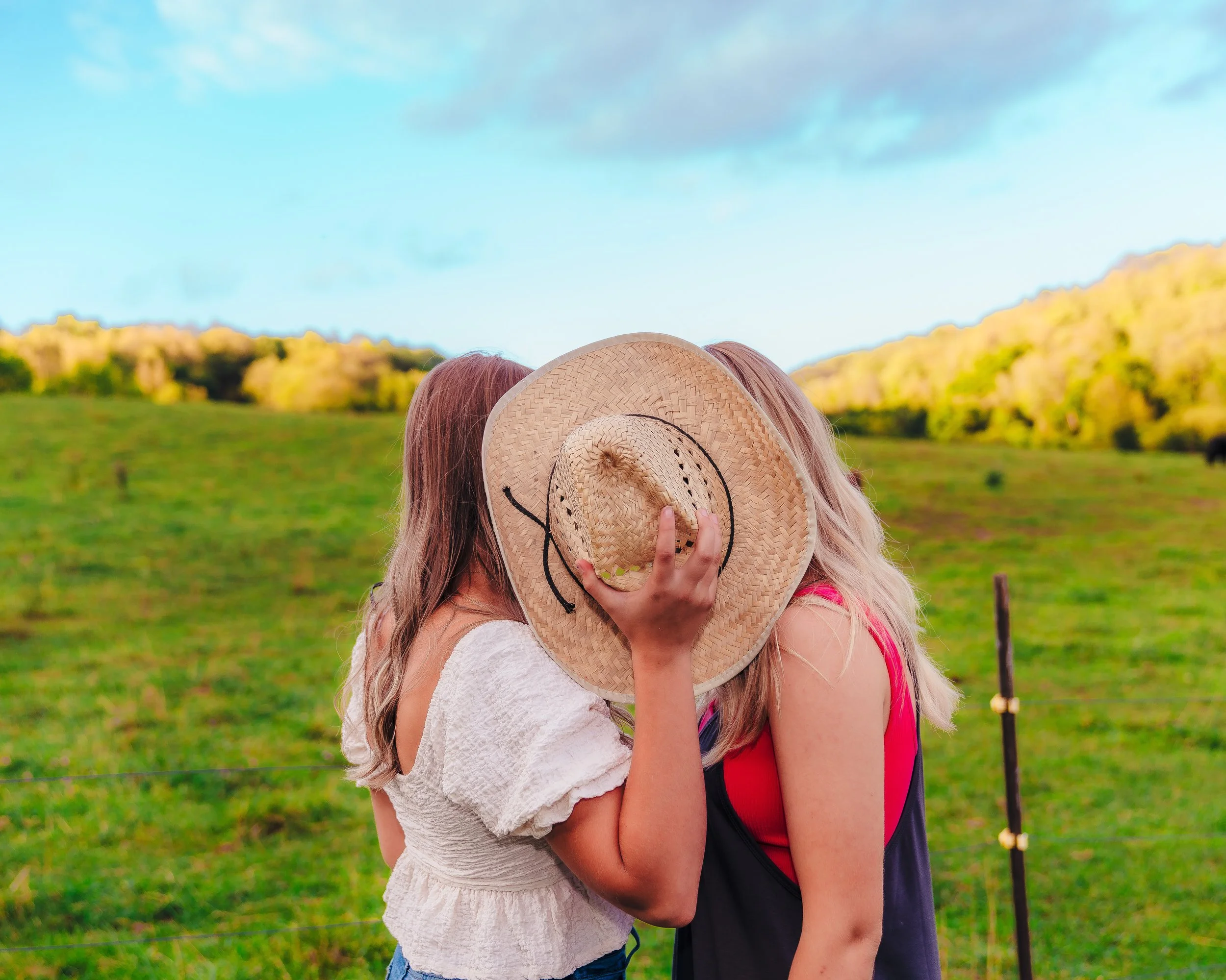 Two women with long hair holding a large straw sunhat, covering their faces, standing in a green open field with trees and hills in the background on a sunny day.