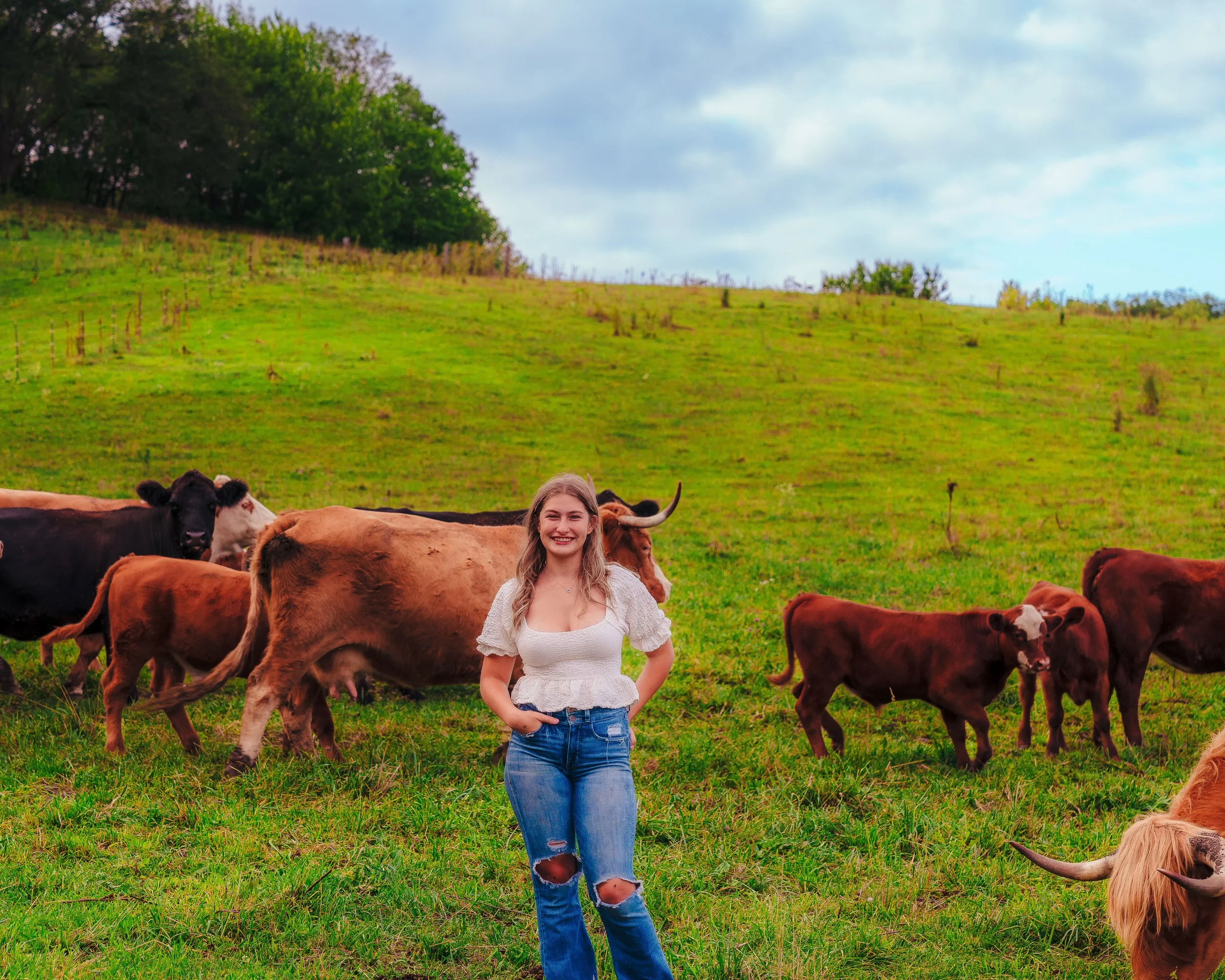 A young woman standing in a grassy field with cows grazing around her on a cloudy day.
