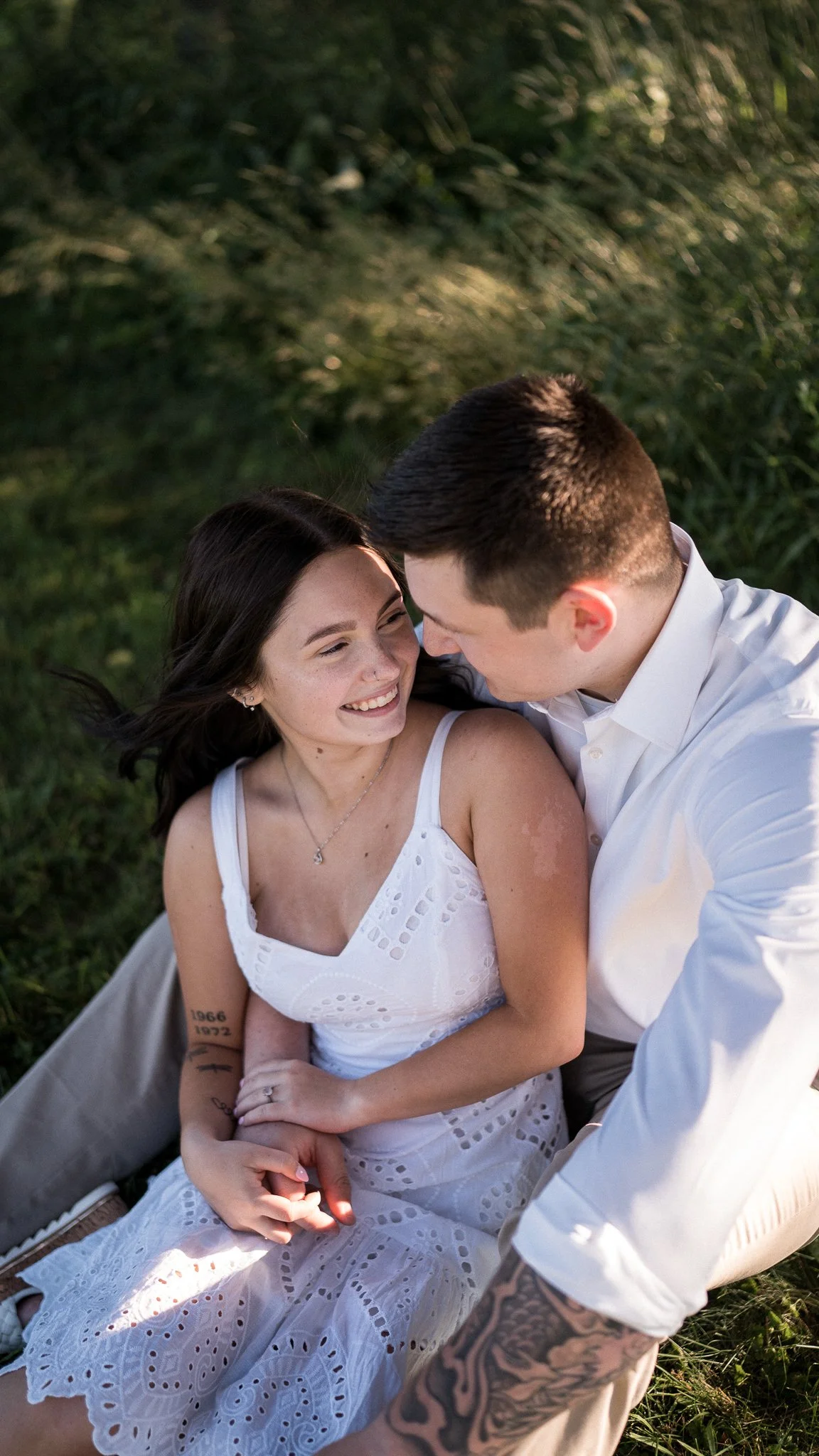 A smiling woman and a man sharing a close moment outdoors, sitting on grass with trees in the background.