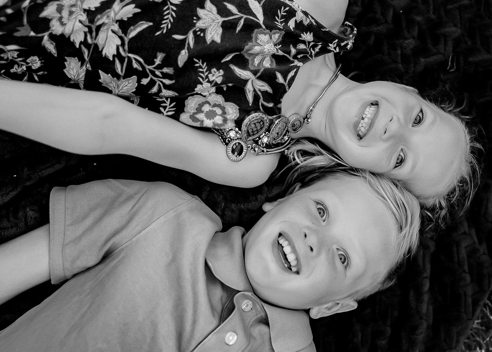 A black-and-white photograph of a smiling woman and a young boy lying on their backs on a dark textured surface. The woman has light-colored hair, wears a floral patterned dress, and a decorative necklace. The boy has short light-colored hair, wears 