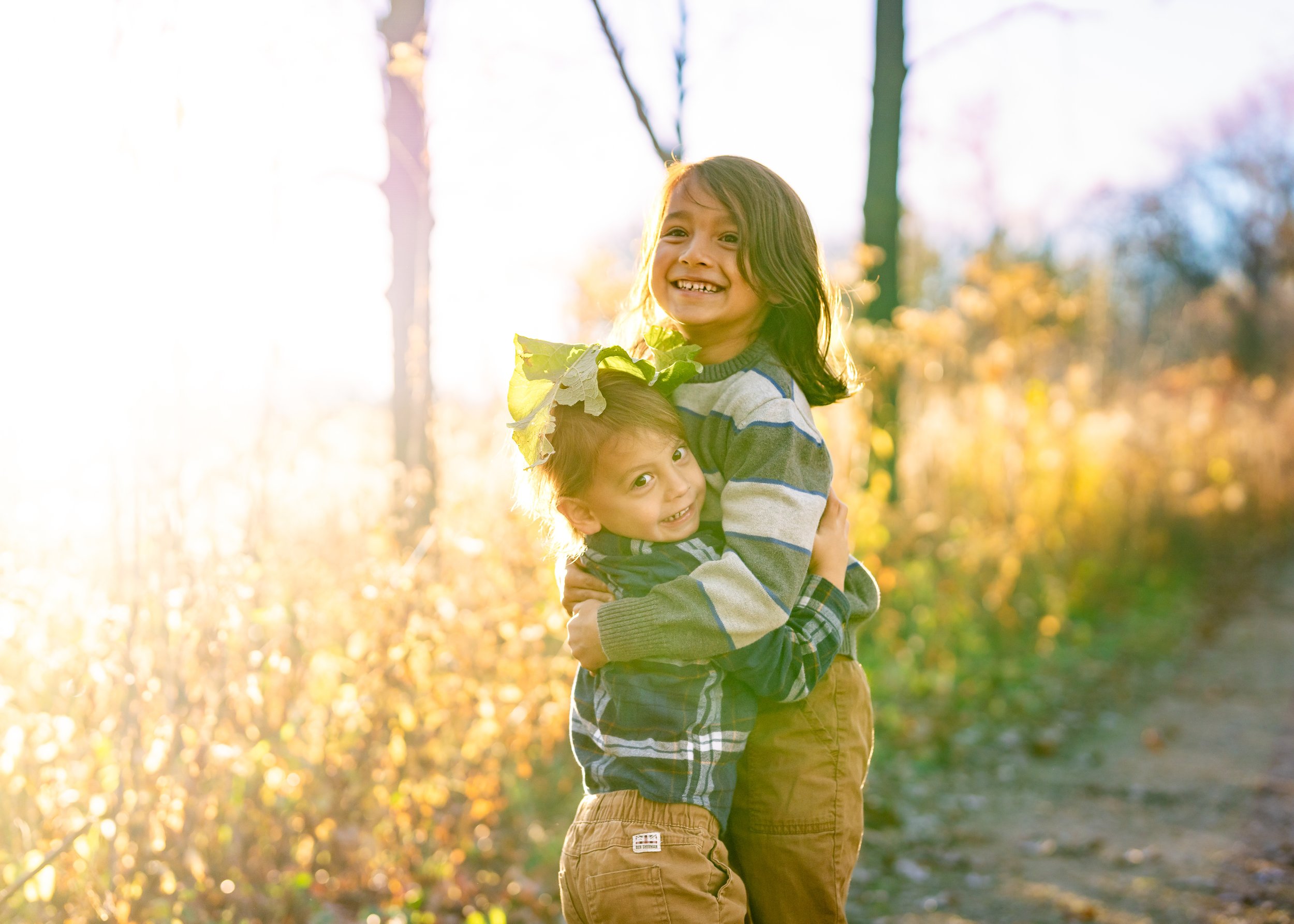 Two children hugging outdoors in a park during autumn, with sunlight shining through trees in the background. The older girl has dark hair and is smiling, while the younger boy has a leaf crown on his head and is also smiling.