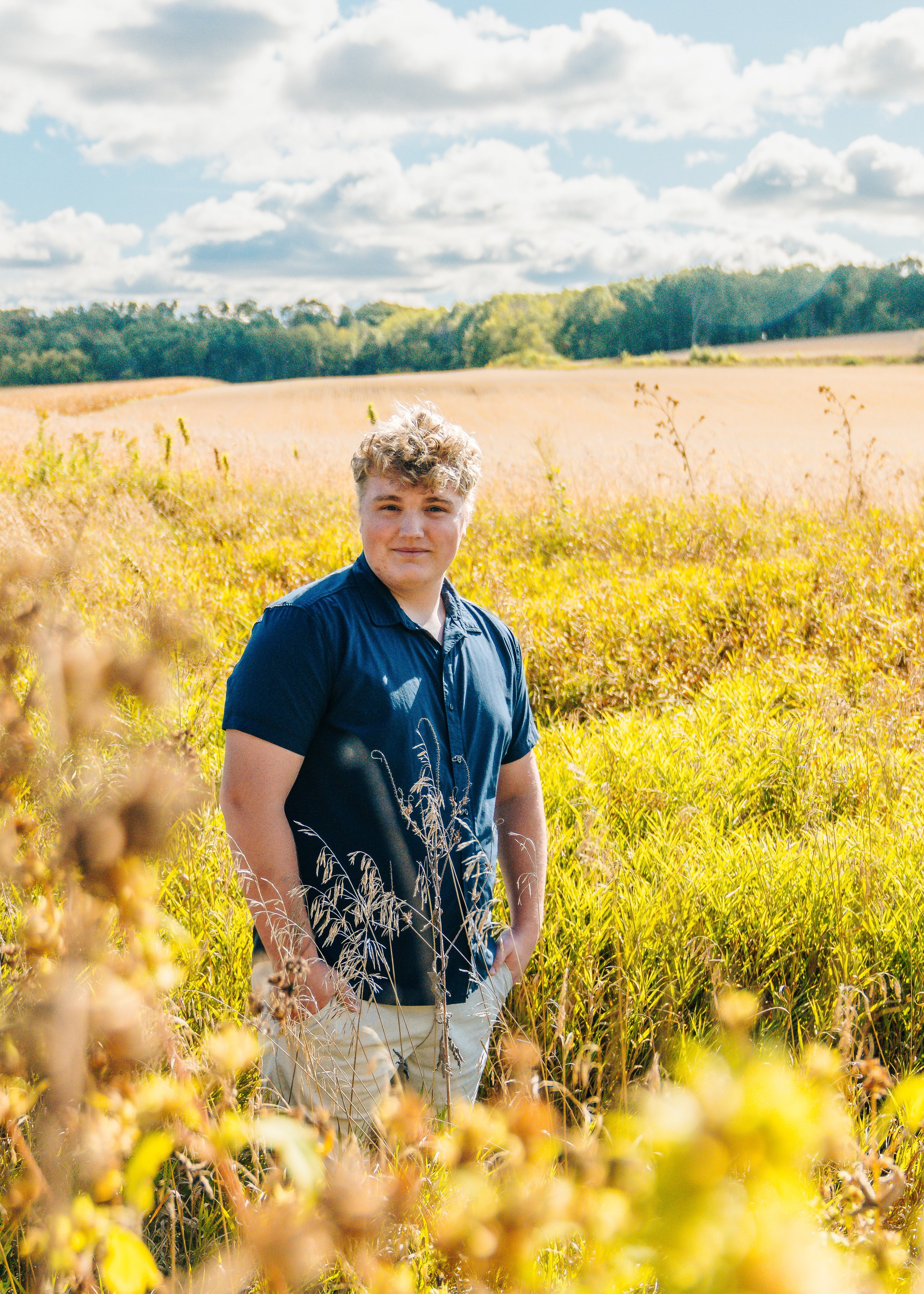 A young man with curly blond hair stands in a yellow and green field on a sunny day with scattered clouds.