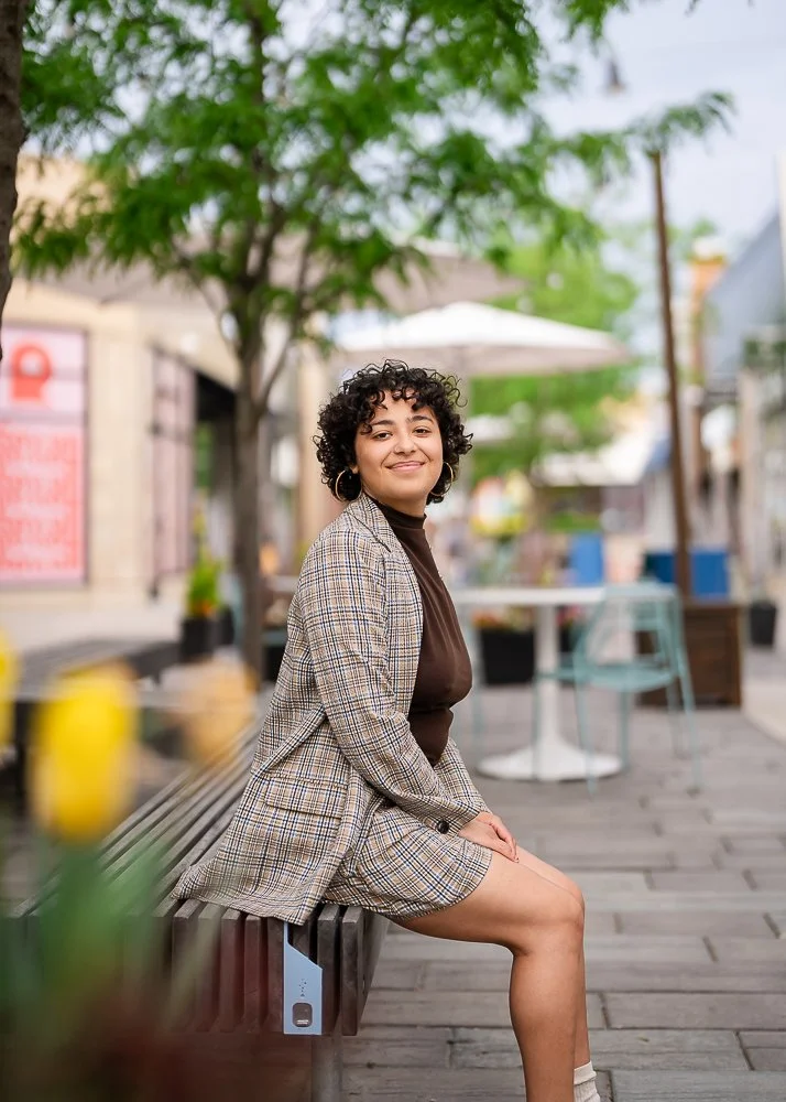 A young woman with curly hair smiling, sitting on a bench outdoors in a city setting with trees, buildings, and tables in the background.