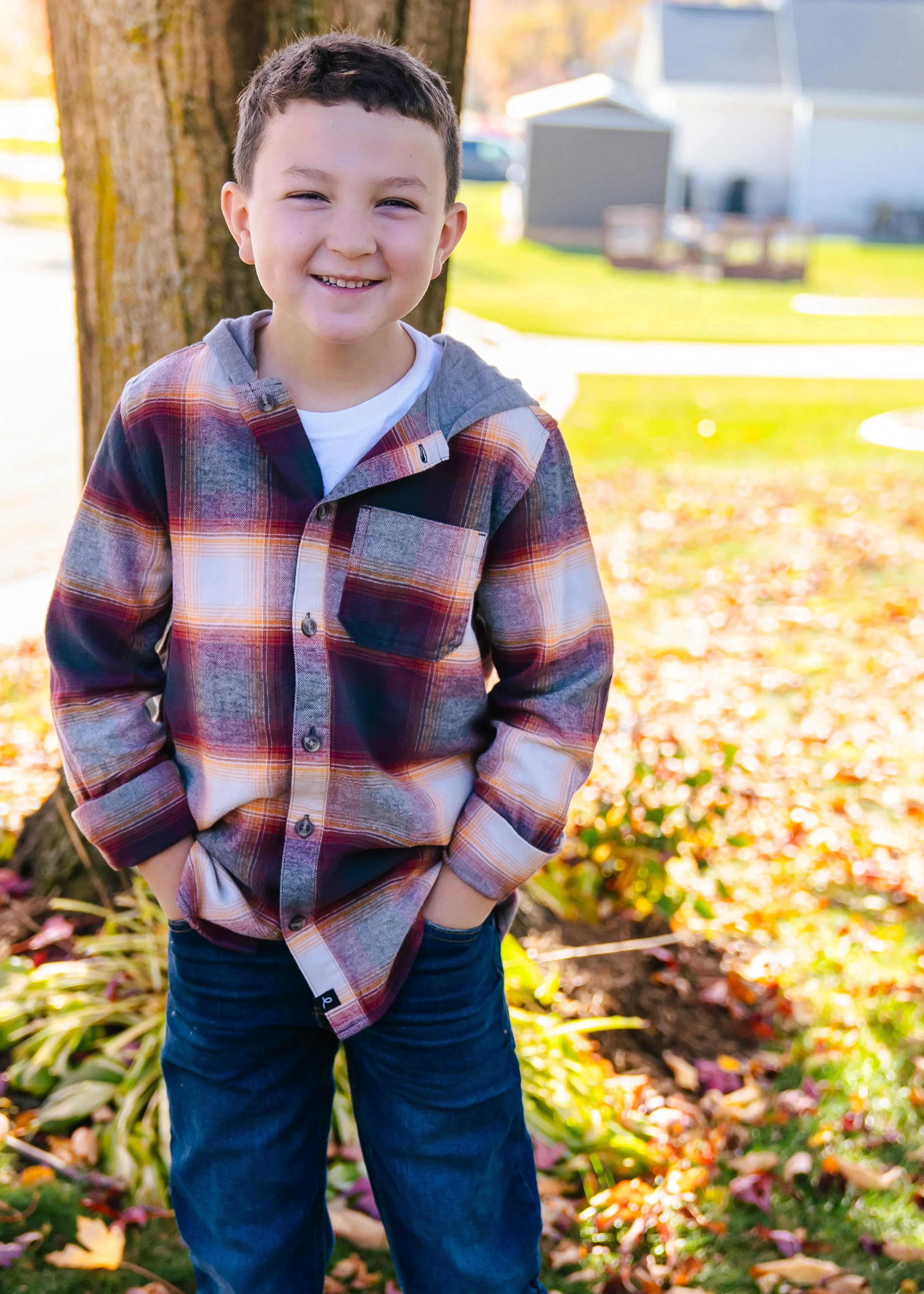 A young boy stands outdoors near a tree, smiling at the camera, with fallen autumn leaves on the ground and houses in the background.