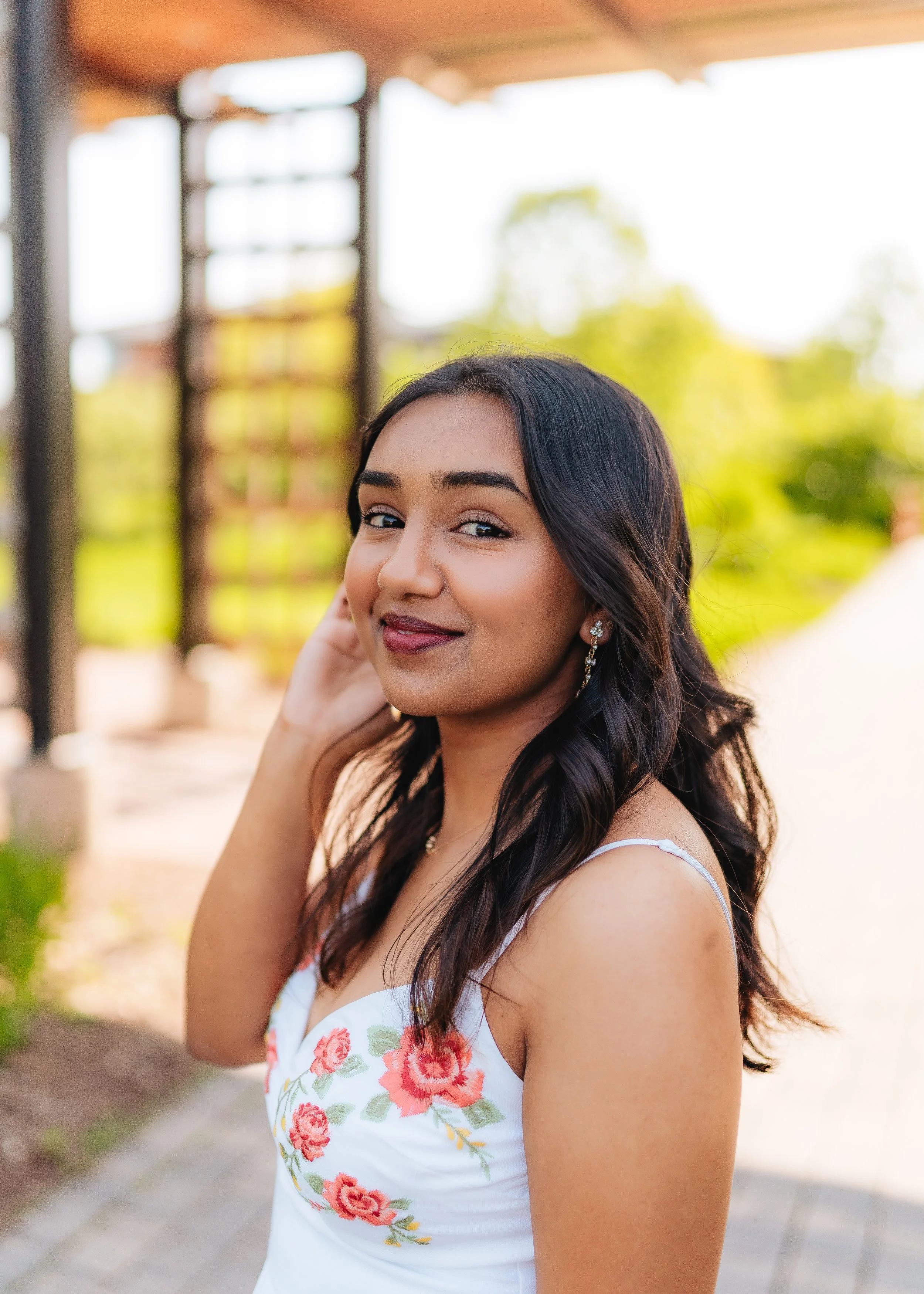 A young woman with long dark hair styled in loose waves, wearing a white dress with floral embroidery, standing outdoors with a blurred background of greenery and wooden structure, smiling softly at the camera.