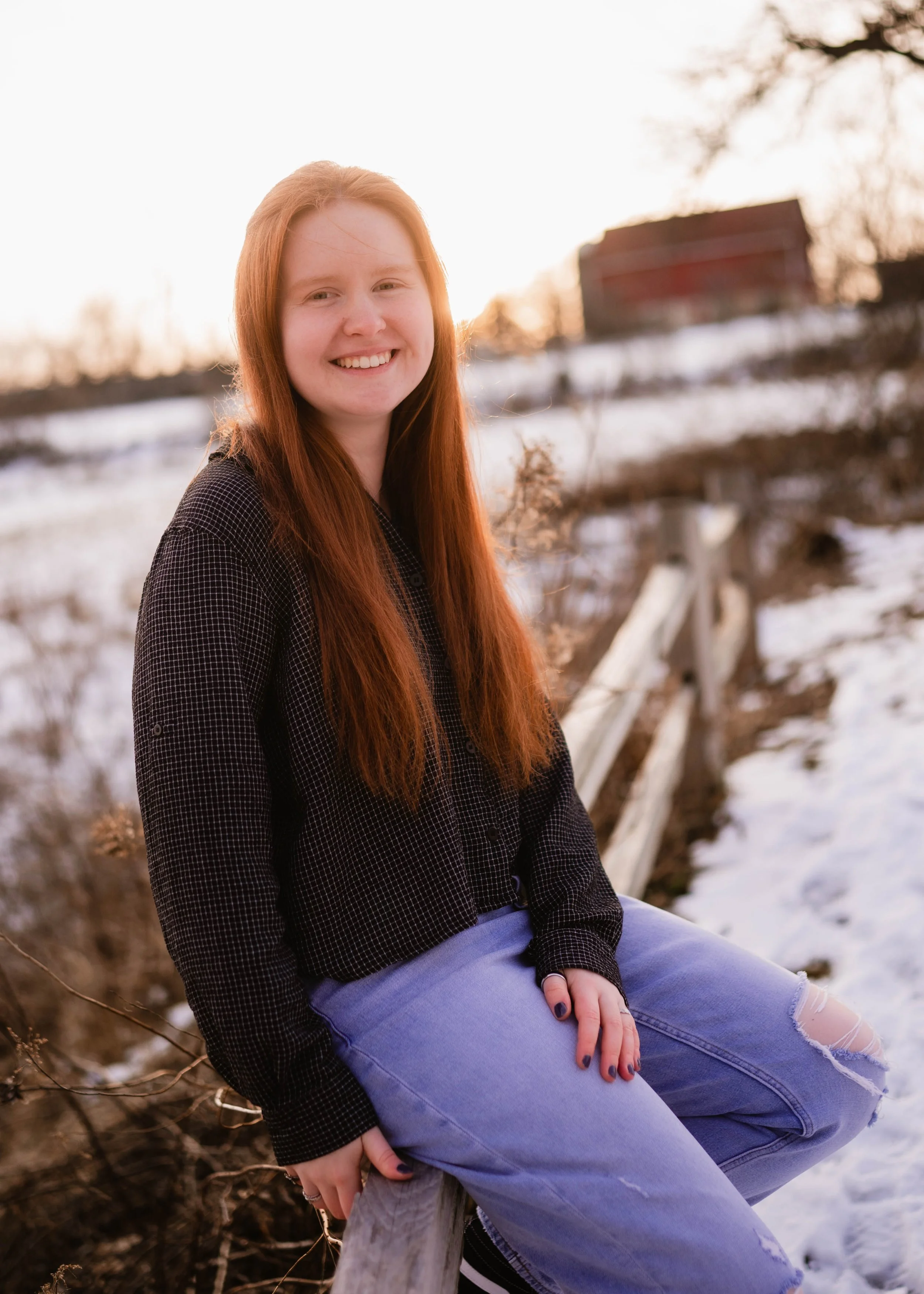 A young woman with long red hair sitting on a wooden fence outdoors during winter sunset, smiling at the camera, wearing a black checkered shirt and ripped blue jeans.