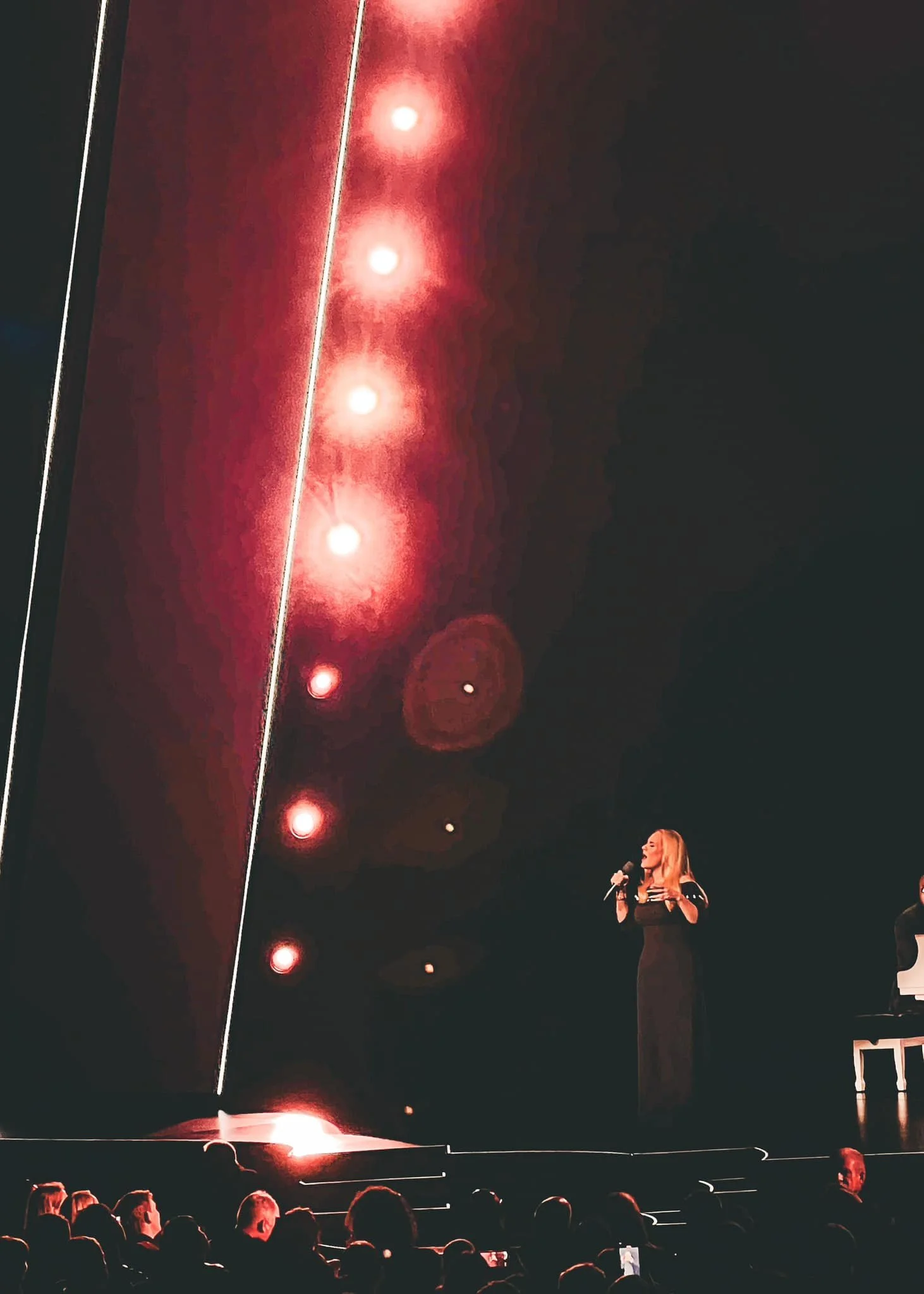 A woman wearing a black dress is singing into a microphone on a stage with a keyboardist beside her. The stage is dark with red lights creating a dramatic atmosphere, and there is a crowd of people watching in the foreground.