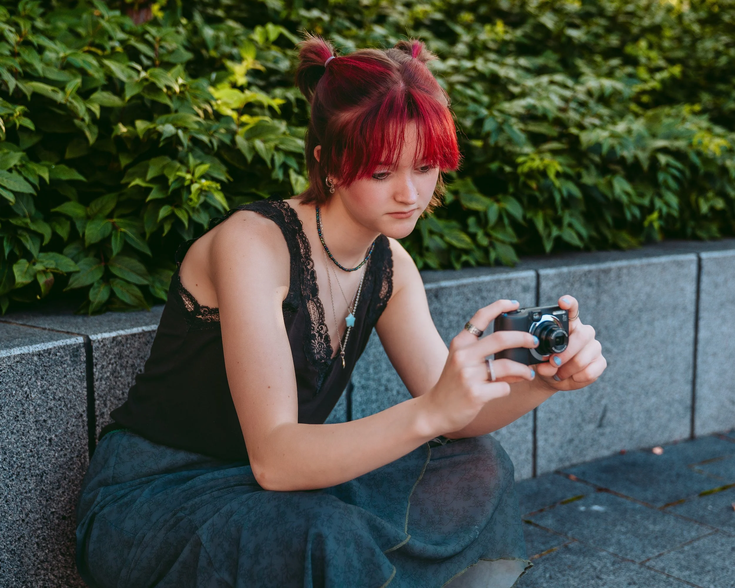 A young woman with short red hair, wearing a black lace top and patterned skirt, sitting on a stone bench outdoors, looking at a camera she is holding.