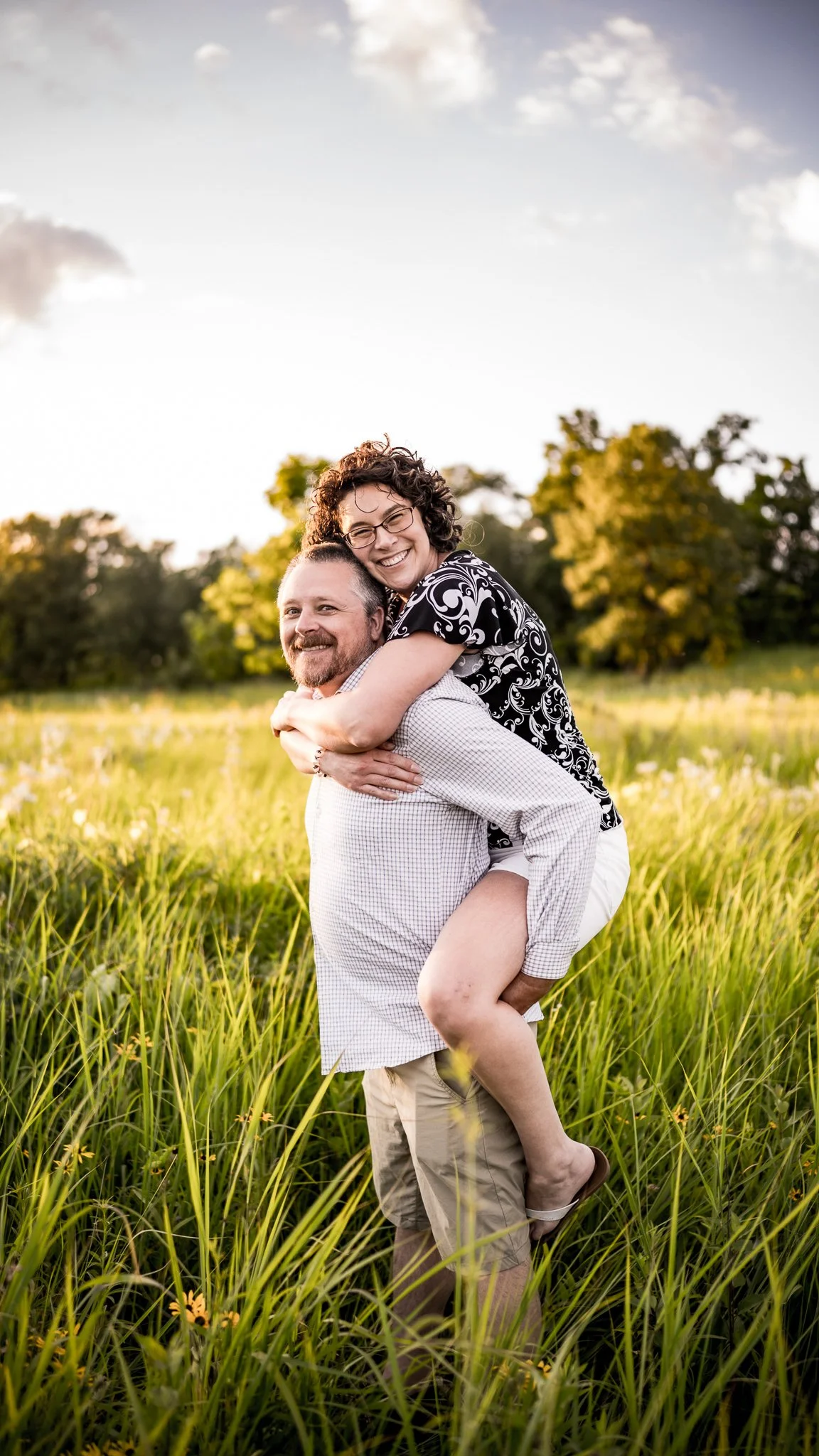 A man giving a piggyback ride to a woman in a green field with tall grass and trees in the background.