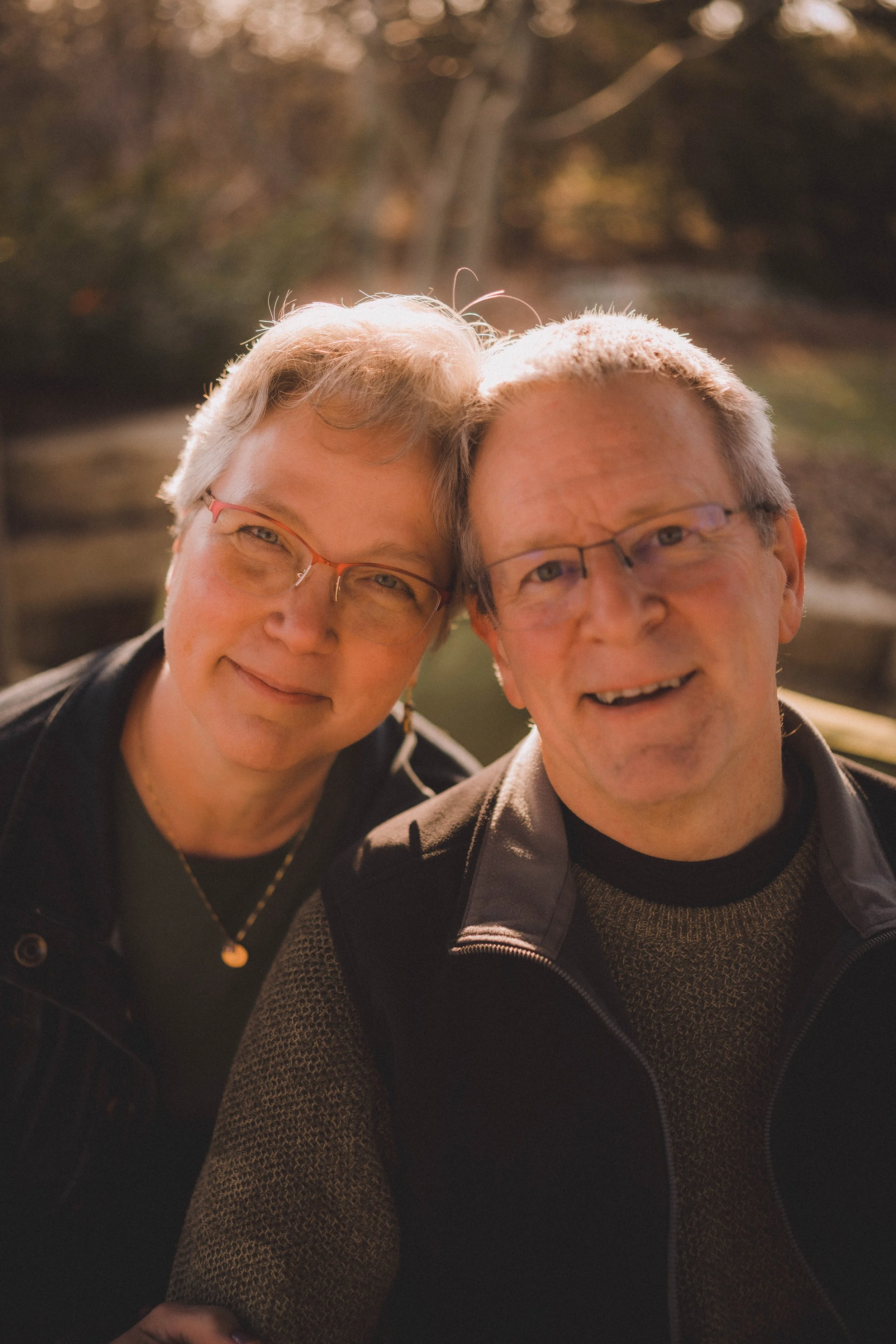 Close-up of an older couple smiling outdoors with trees and sunlight in the background.