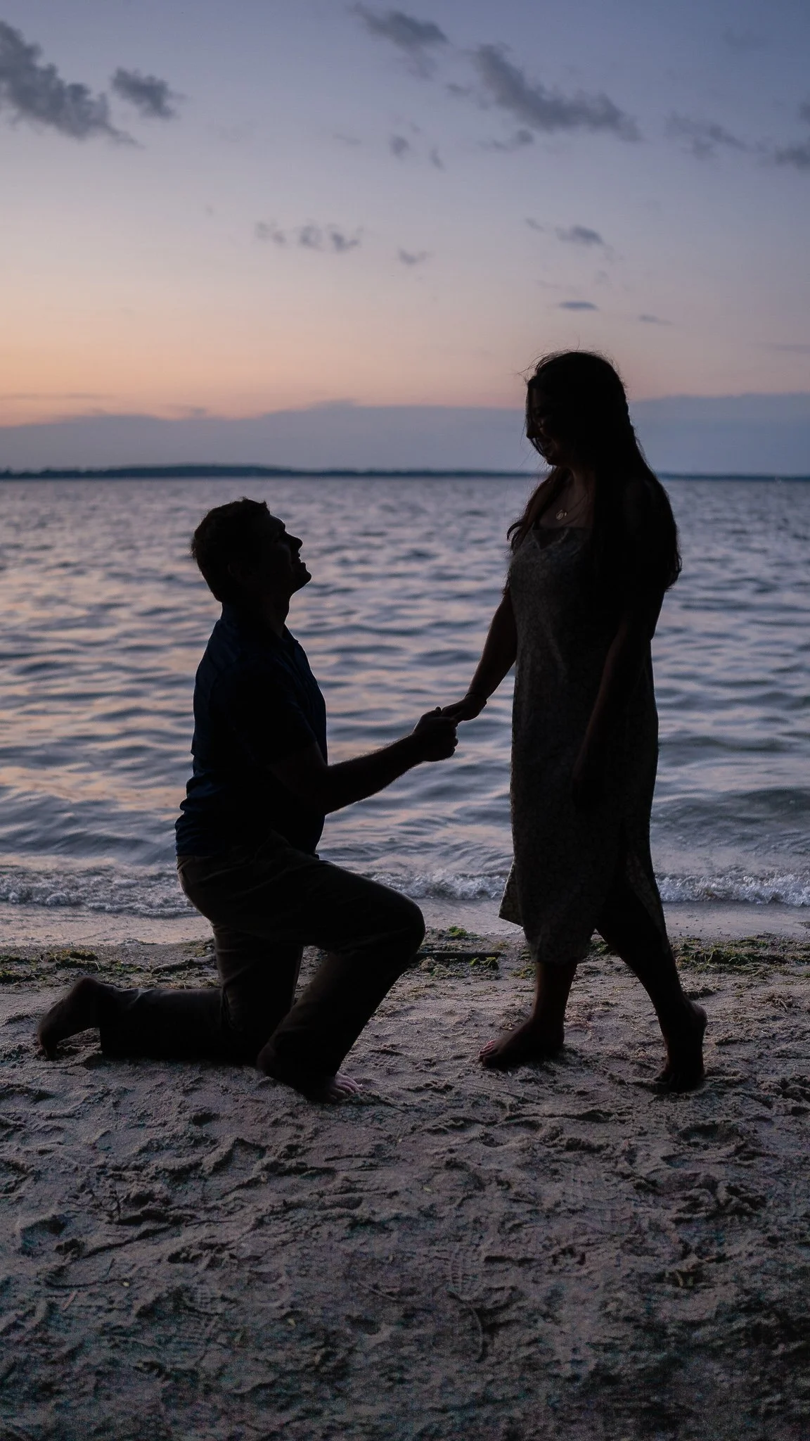 A silhouette of a man kneeling on one knee and holding the hand of a woman standing on the beach during sunset, with the ocean in the background.