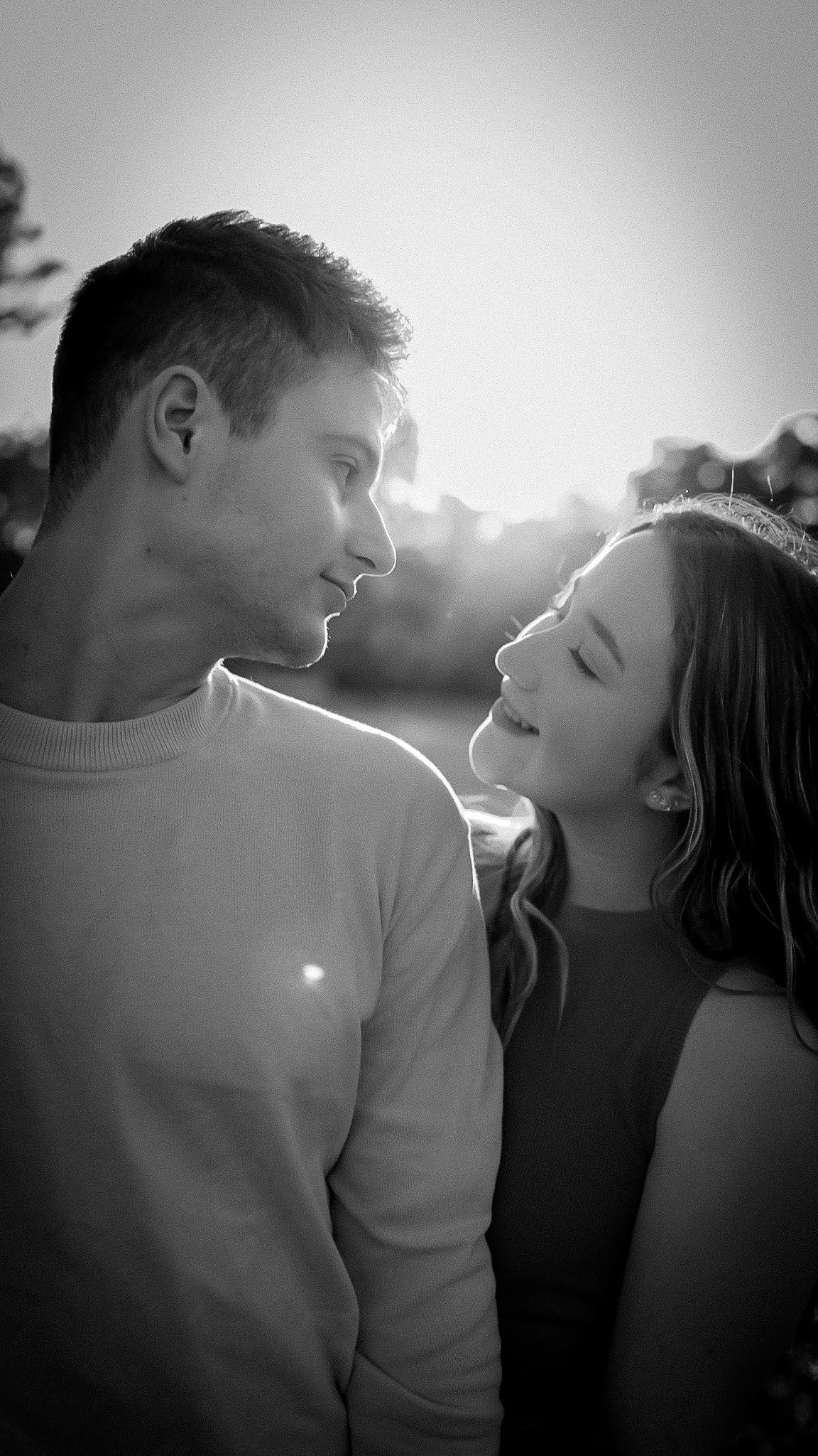 A black and white photo of a young man and woman gazing at each other, with the sunlight behind them creating a backlit effect.
