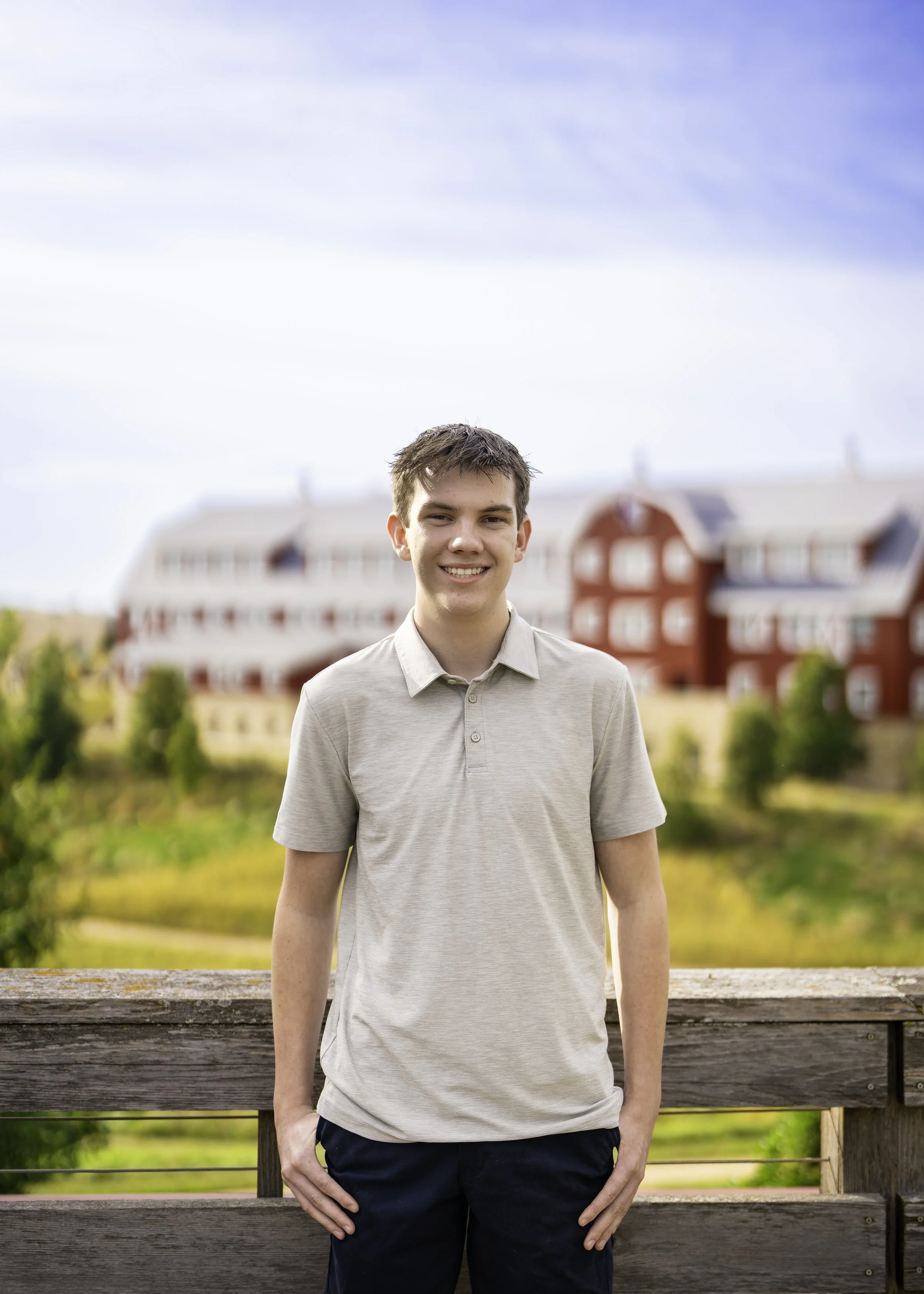 A young man standing outdoors on a wooden railing, smiling, with red and white buildings and greenery in the background under a partly cloudy sky.