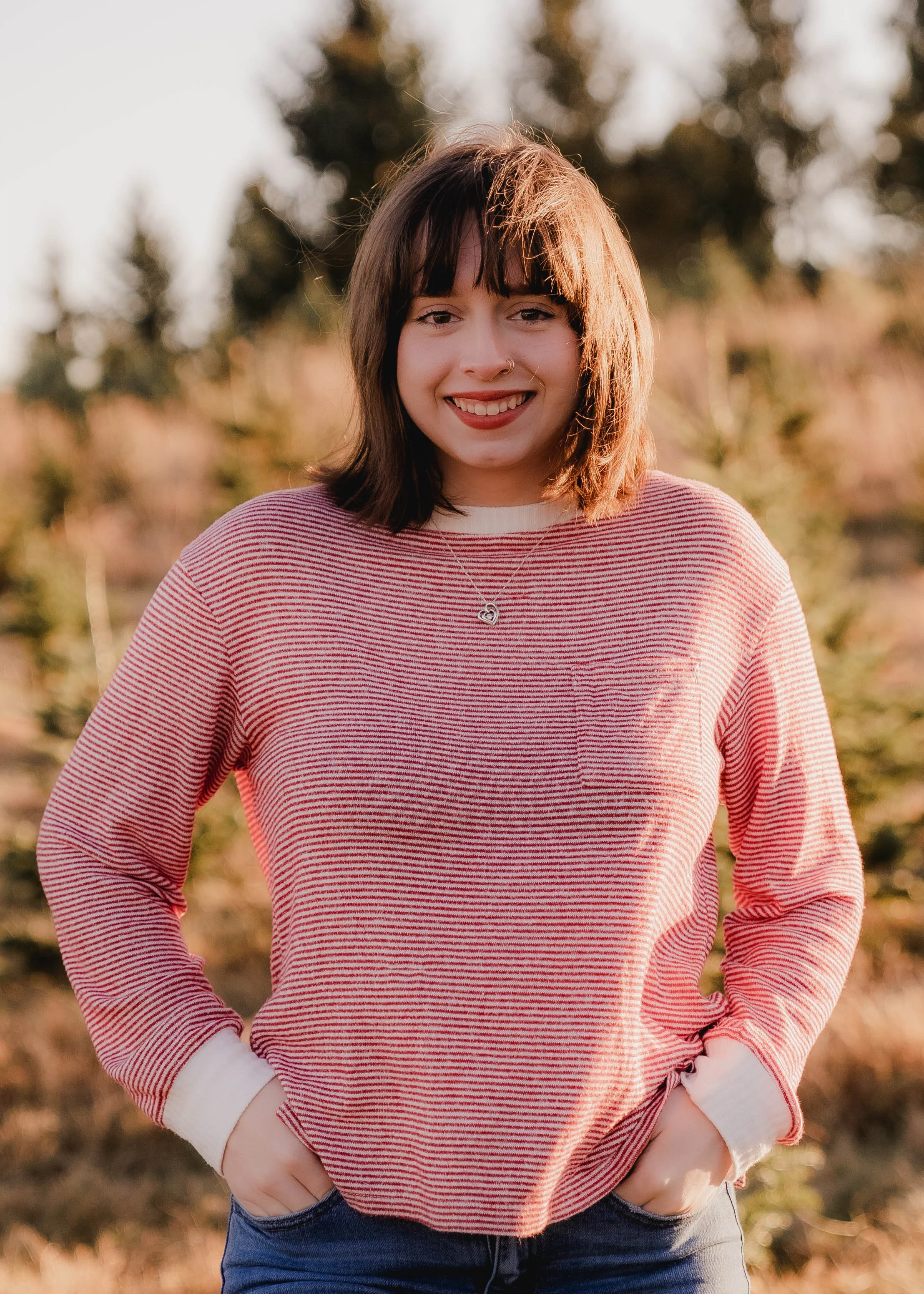 A young woman with shoulder-length dark hair, wearing a red and white striped long-sleeve shirt, standing outdoors with a blurred background of trees, smiling at the camera, during sunset.