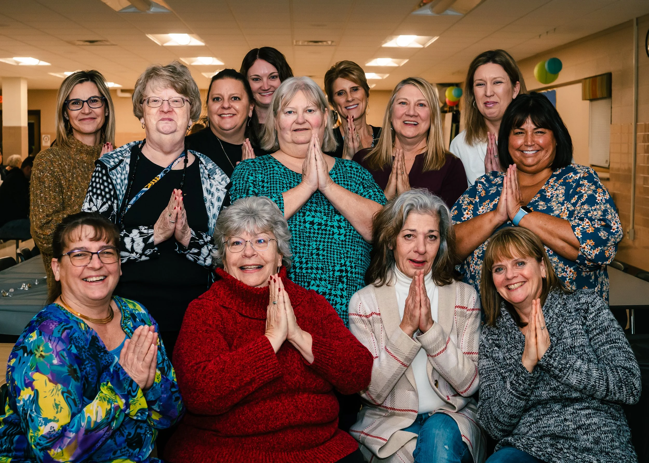 A group of smiling women, some with hands pressed together in a prayer-like gesture, gathered indoors in a celebratory or spiritual setting.