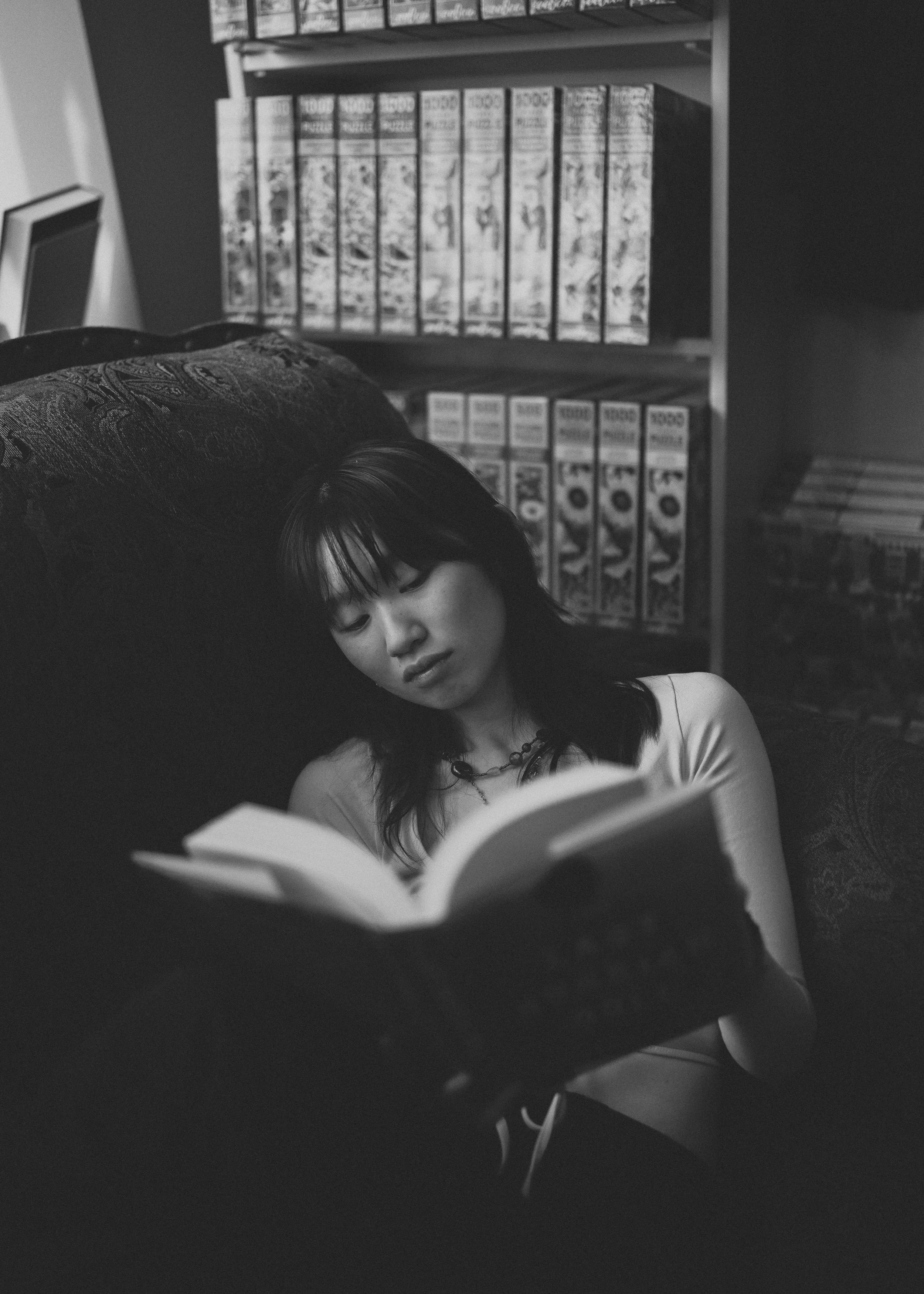 A woman with dark hair and bangs reading a book and resting against a dark pillow in a room with a shelf full of comic books or vintage boxes in the background.