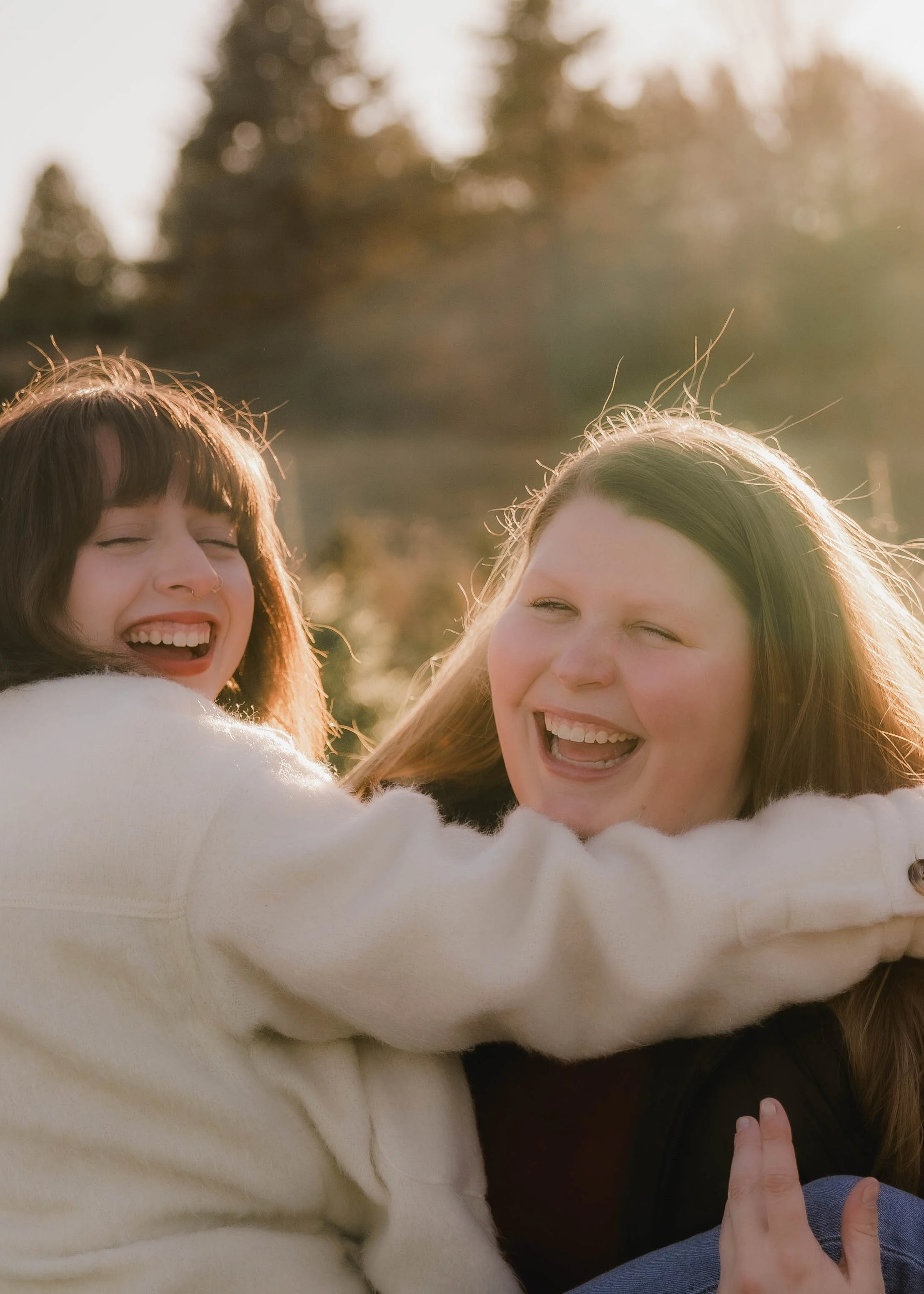 Two young women laughing and hugging outdoors during golden hour with sunlight behind them and trees in the background.