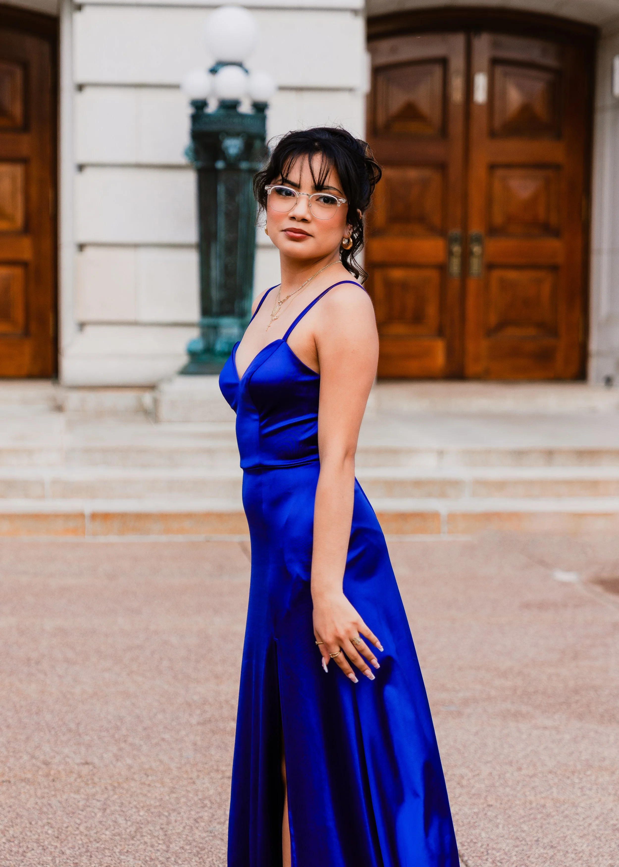 A young woman wearing a royal blue satin evening gown with thin straps, standing outdoors in front of a building with wooden doors. She has dark hair styled in loose waves, is wearing glasses, and accessorized with jewelry.