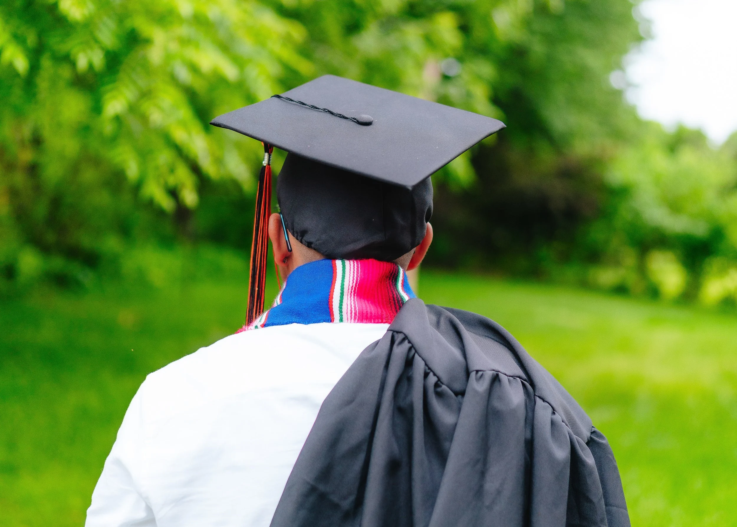 Person in graduation cap and gown walking outdoors on a grassy path with green trees in the background.