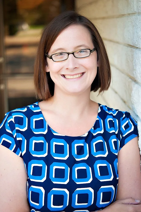 Woman with short brown hair and glasses smiling for a photo wearing a blue dress