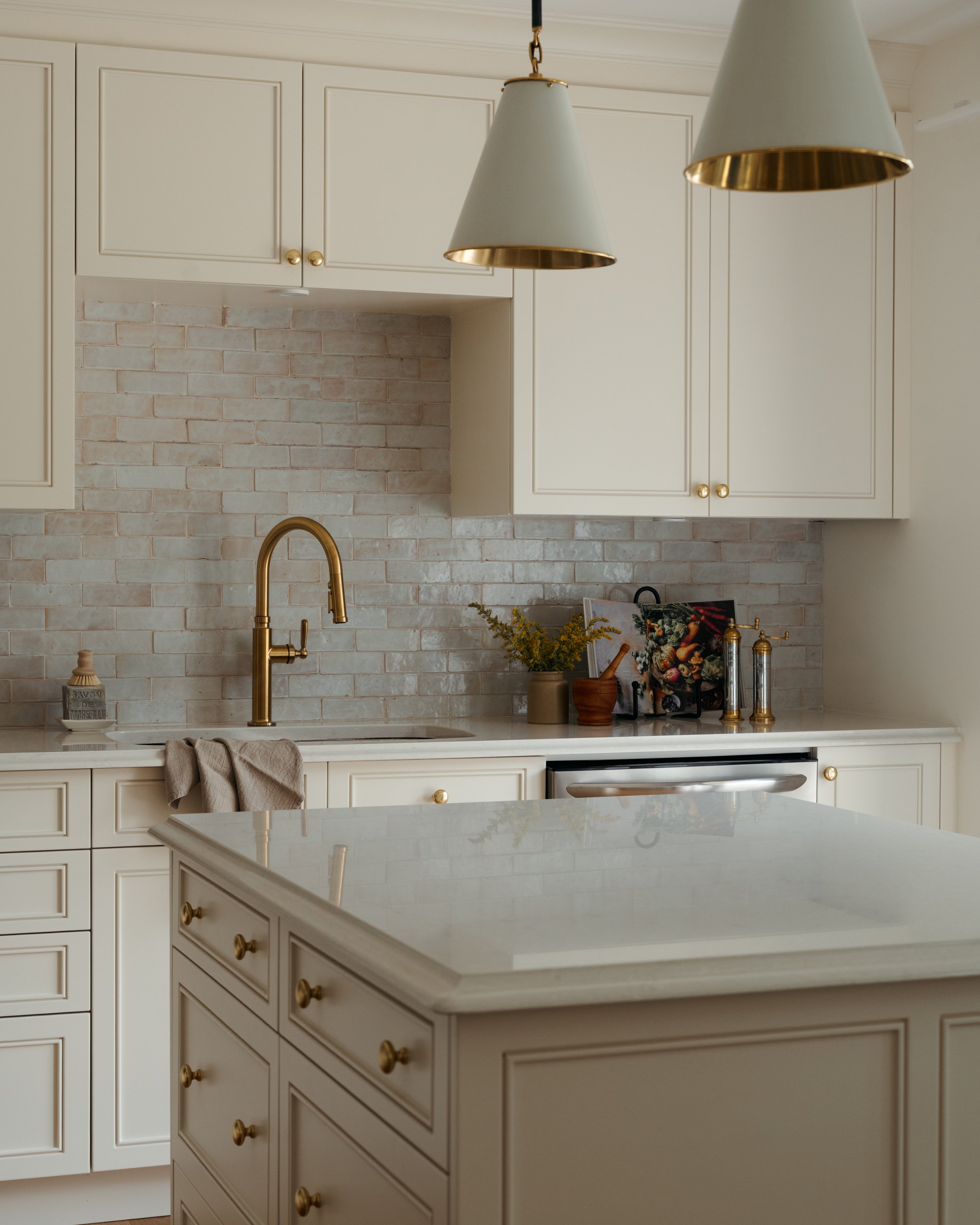 Candace Plotz Design's Project Renewed Roots showcases a luminous kitchen with soft ivory cabinetry, brass knob hardware, and honed white marble countertops. Handmade terracotta brick backsplash tiles create warmth beneath white shaker-style upper ca