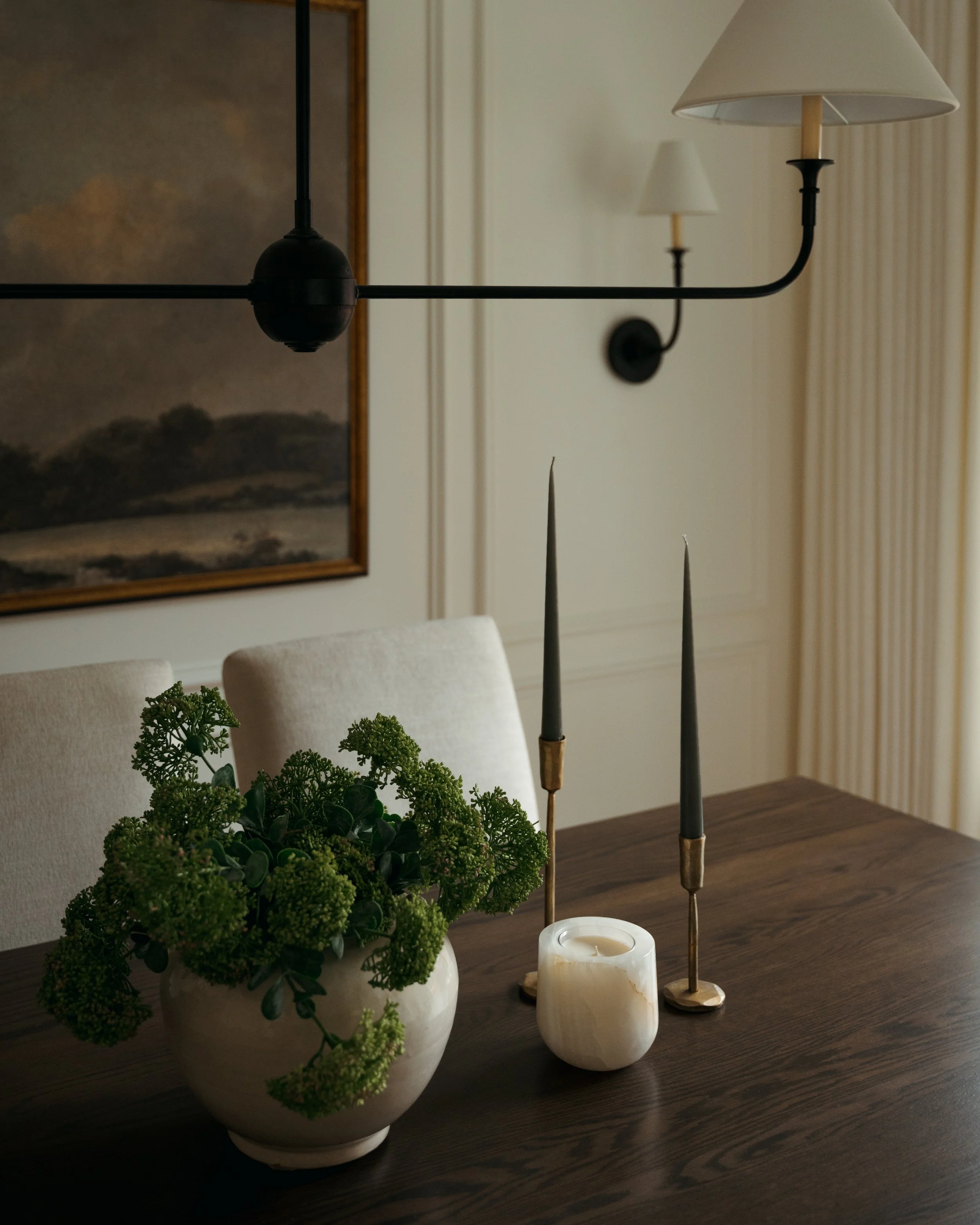 Candace Plotz Design's Project Renewed Roots showcases an intimate dining table vignette with golden hour lighting. A dark wood table surface displays a ceramic bowl filled with fresh green parsley, tall sculptural taper candlesticks in brass and bla